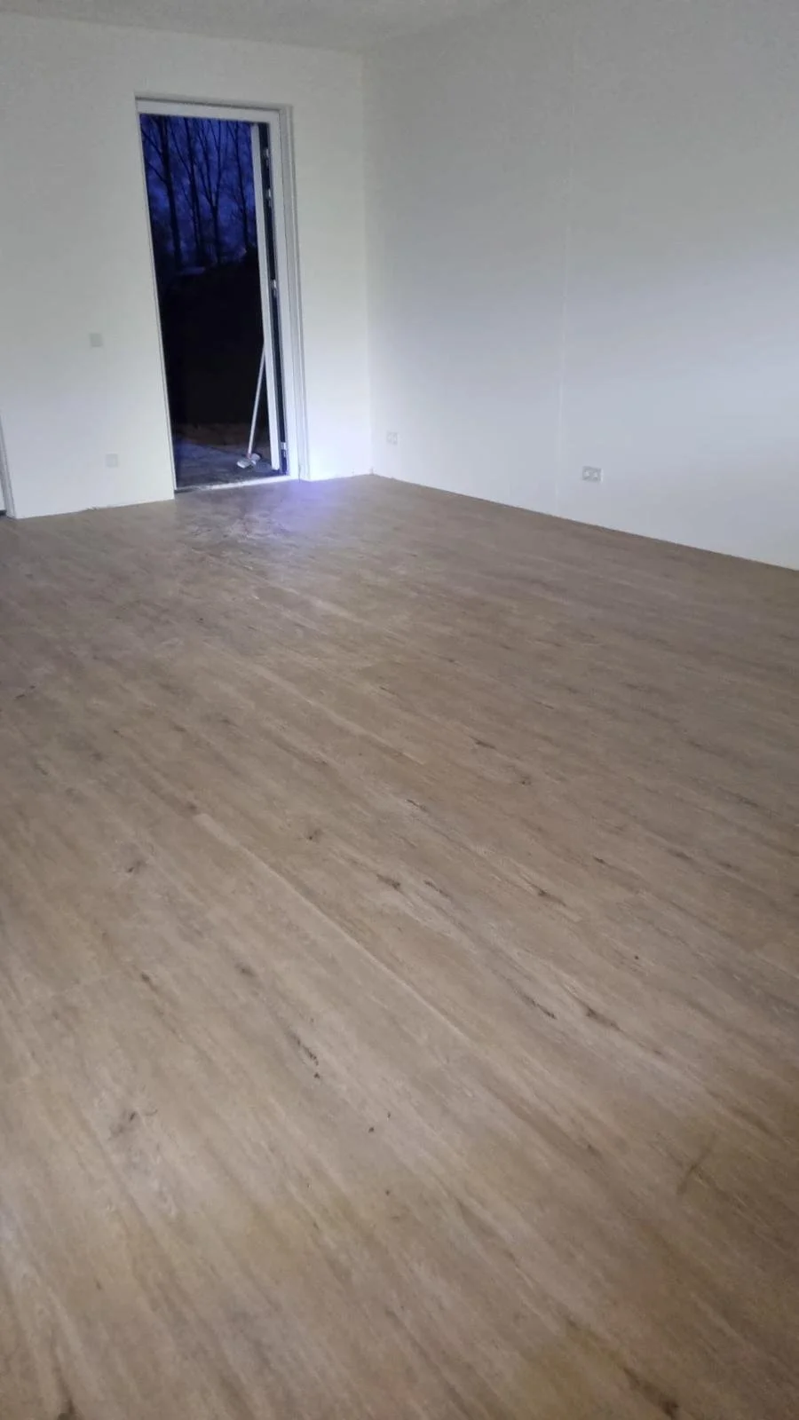 Empty room with newly installed wood flooring, white walls, and a door leading outside at dusk.