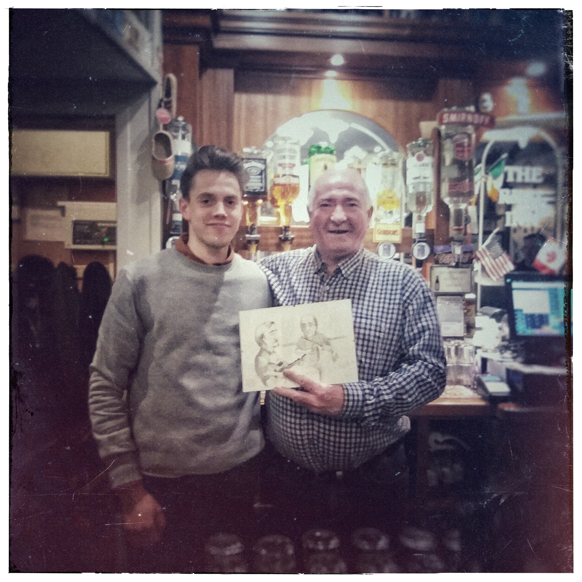 Two men in a bar posing for a photo, with one man holding a drawing of two people. The bar has various bottles and decorations in the background, including flags and signs.