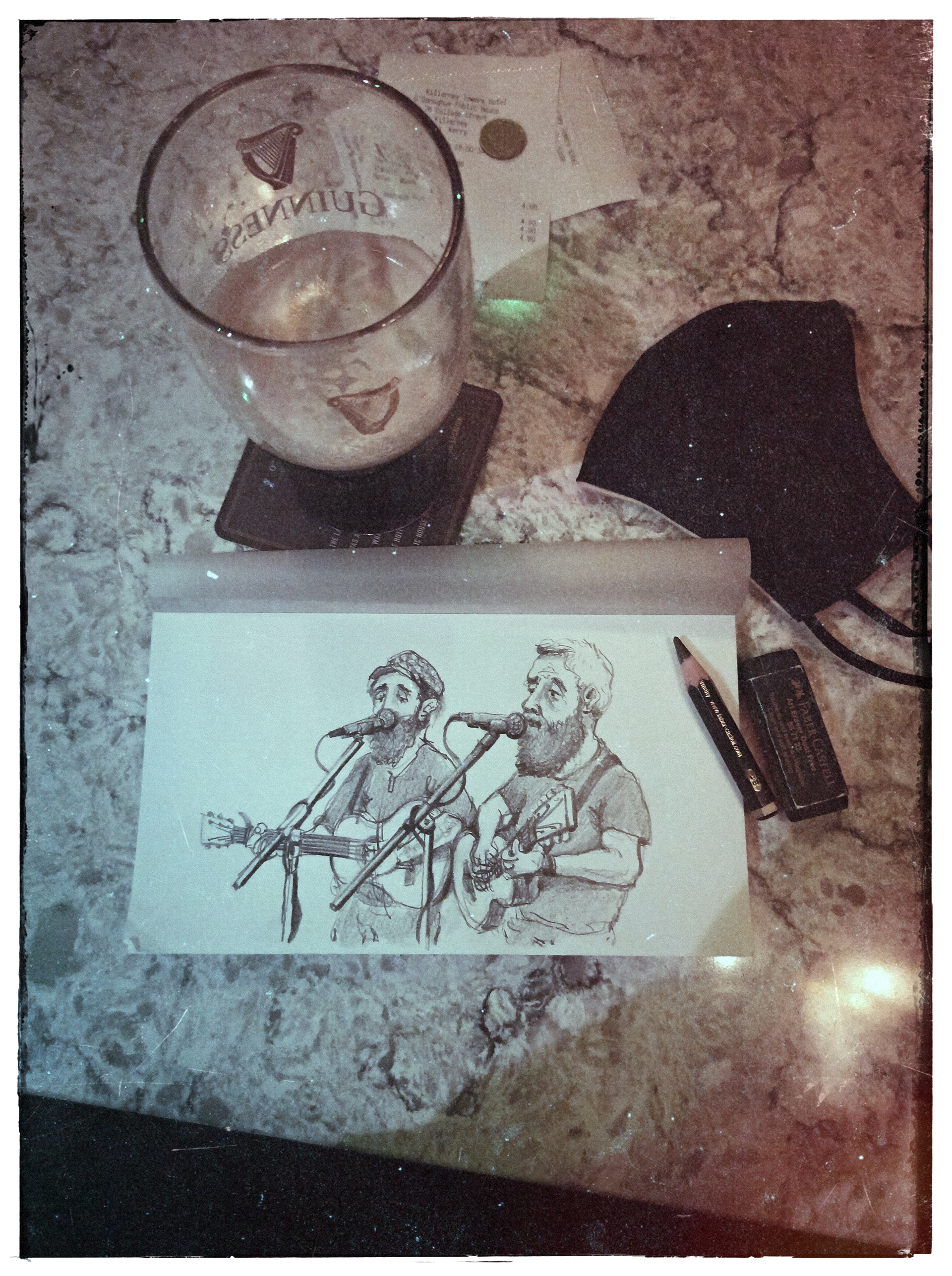 A drawing of two musicians singing into microphones, one playing a guitar, on a marble pub table with a glass of Guinness beer, a face mask, a pen, and an eraser.