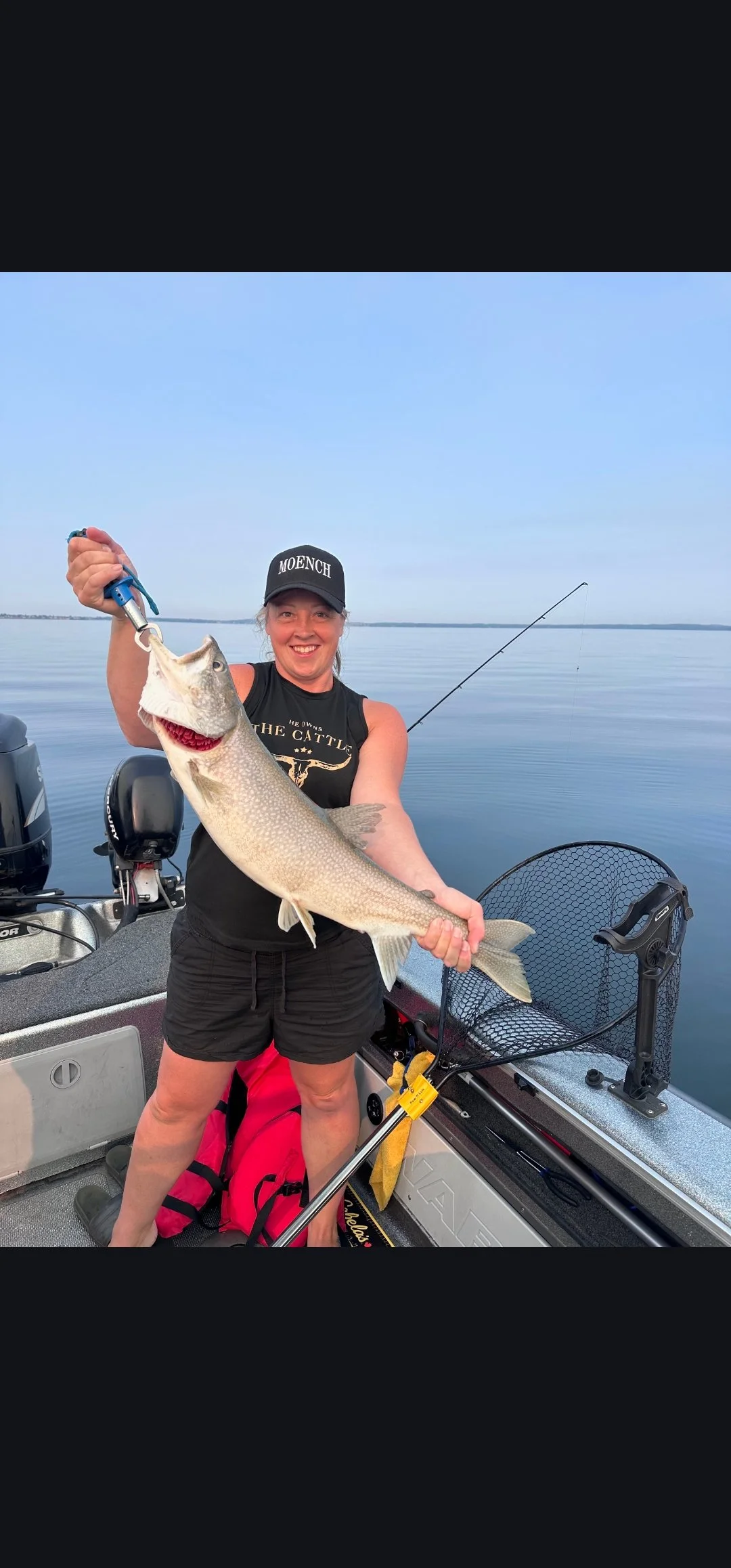 A woman on a boat holding a large lake trout she caught, smiling, with a calm lake and clear sky in the background.