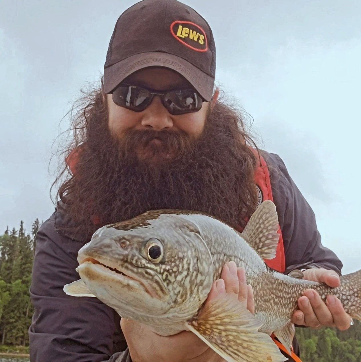 Nature Nathan with a large beard, wearing a baseball cap and sunglasses, holding a lake trout outdoors in nature.