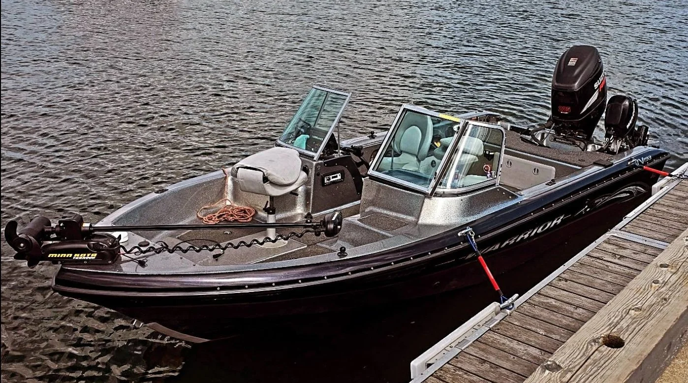 A boat docked at a wooden marina on the water, with an outboard motor, fishing equipment, and two windshielded seats wating for guest to arrive.
