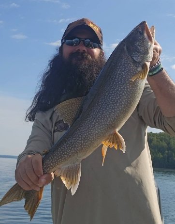 A man with a beard, wearing sunglasses and a cap, holding a large fish outdoors near a body of water.