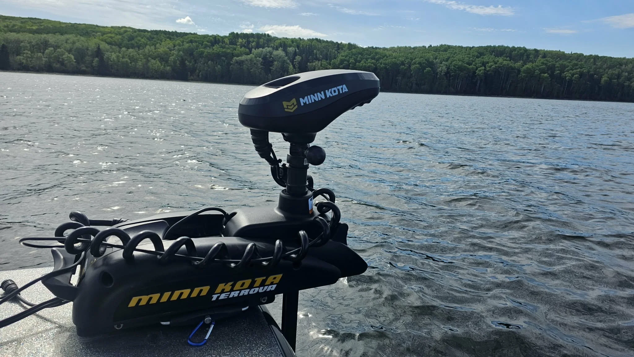 Close-up of Minn Kota trolling motor attached to a boat on a lake with a forested shoreline, under a partly cloudy sky.