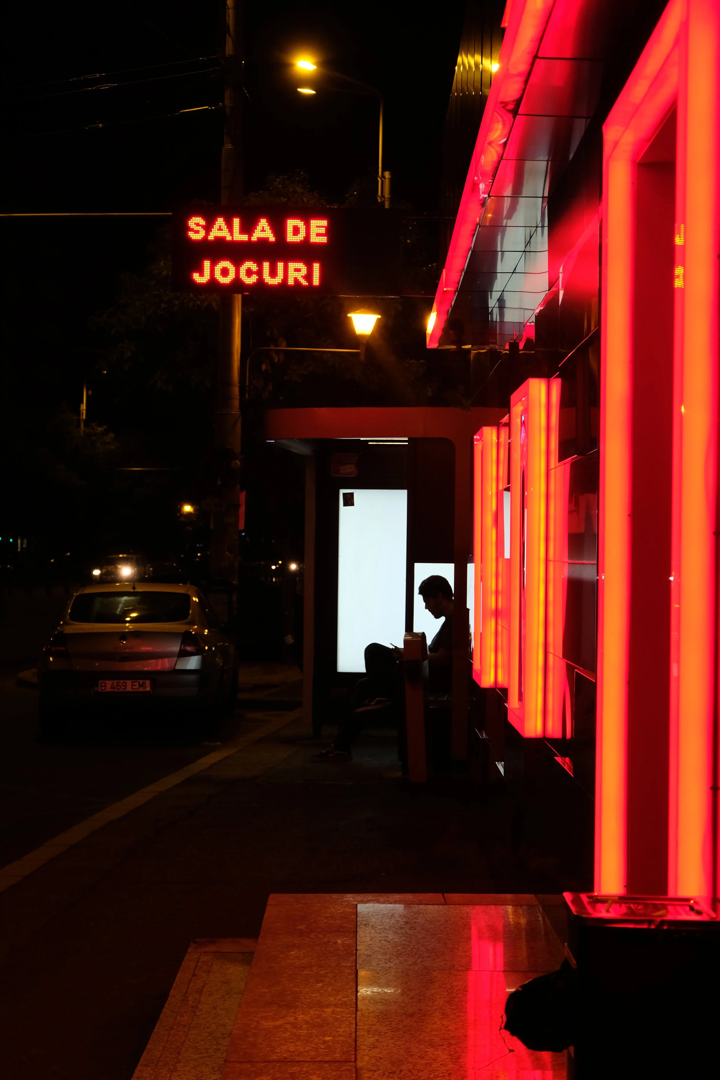 Nighttime scene at a bus stop with a person sitting on a bench and looking at their phone, illuminated by bright red neon lights and a digital sign that says 'SALA DE JOCURI'.
