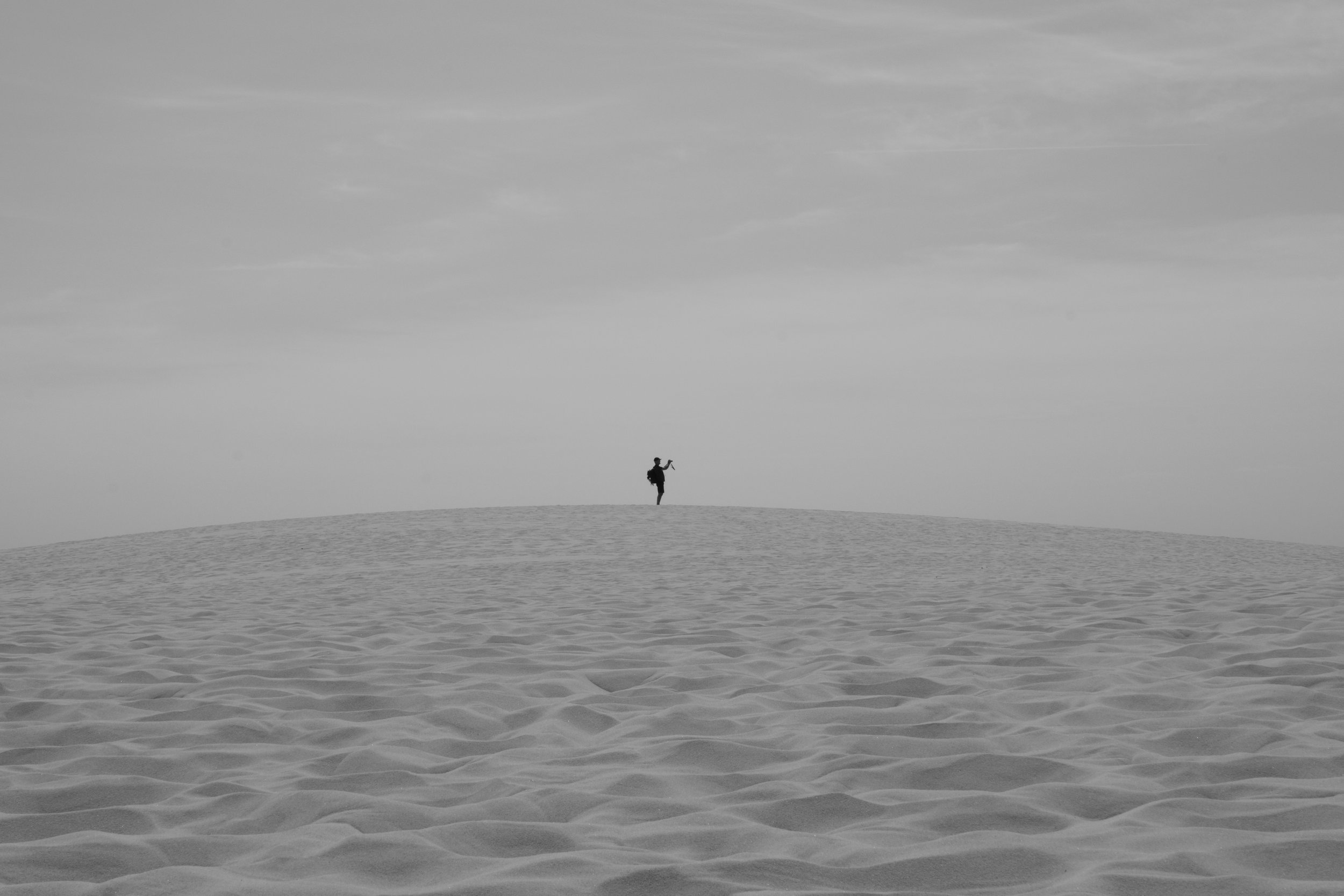 A person standing alone on a sand dune, holding a camera or phone, with a cloudy sky above.