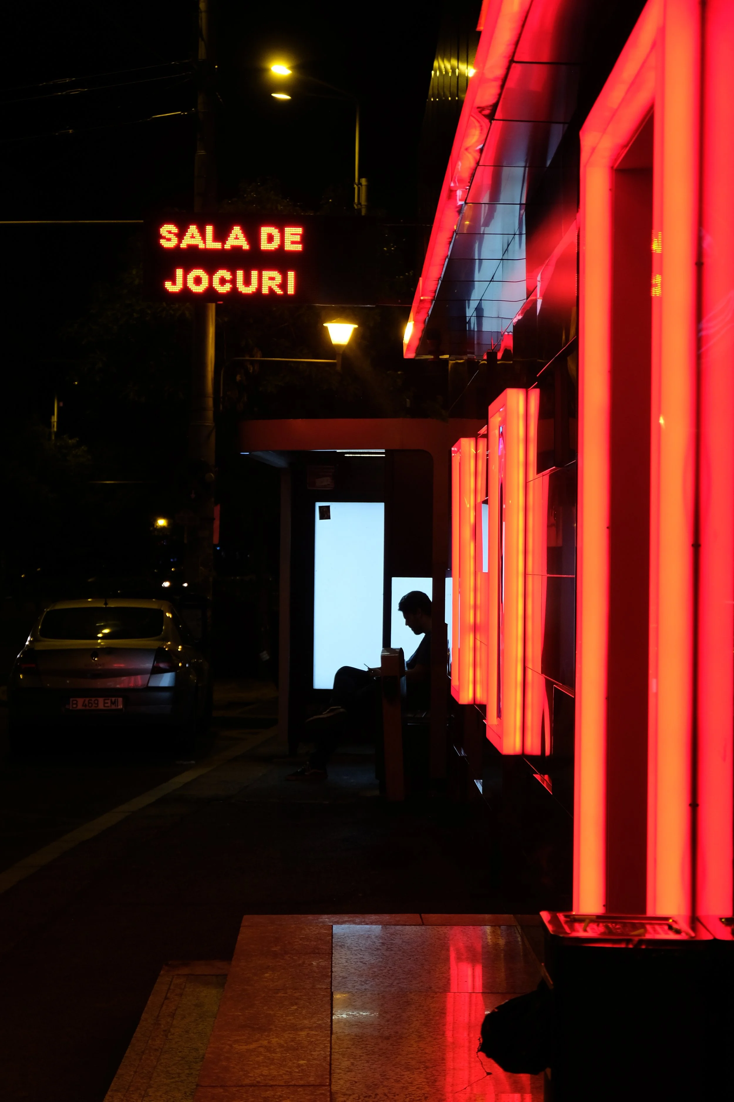 Night scene outside an arcade with a neon sign in Romanian reading "Sala de Jocuri" and a seated person using a phone at a bus stop.