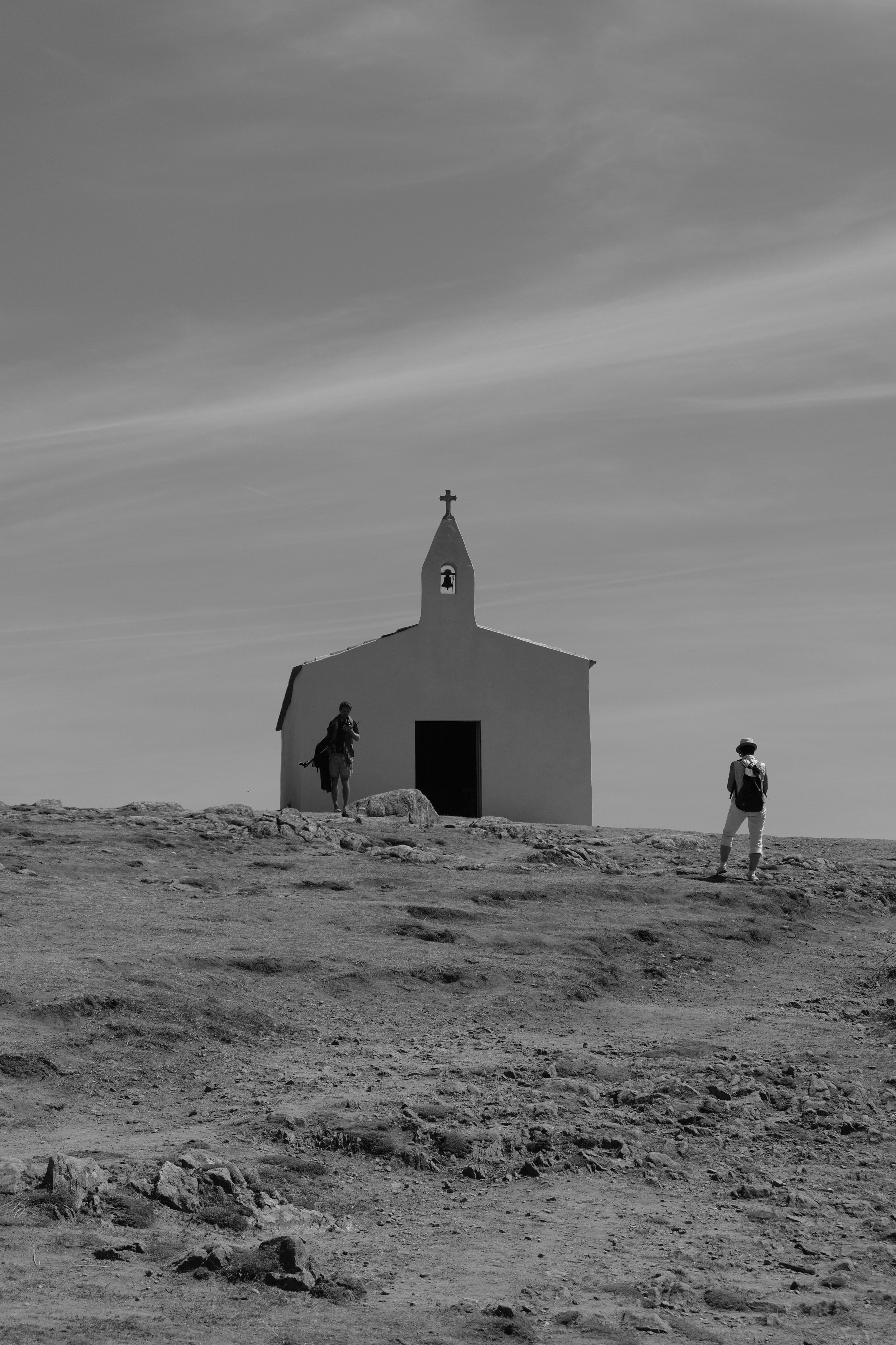A small white church on a hill with a cross on top, two hikers with backpacks approaching it, under a sky with wispy clouds, black and white photo.