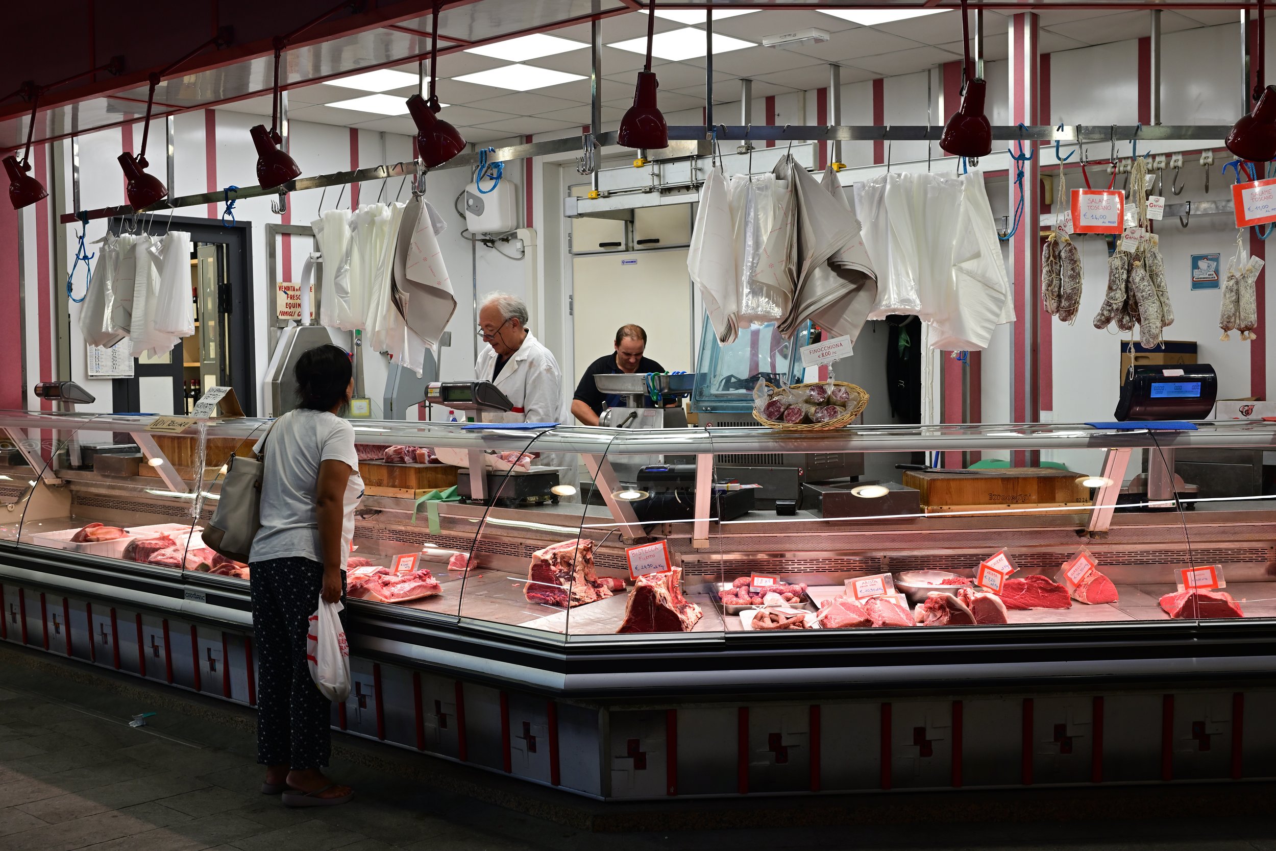 A customer at a meat shop talking to a butcher behind a glass display filled with various cuts of raw meat. The shop has hanging sanitized covers and sausages, with two employees working behind the counter.