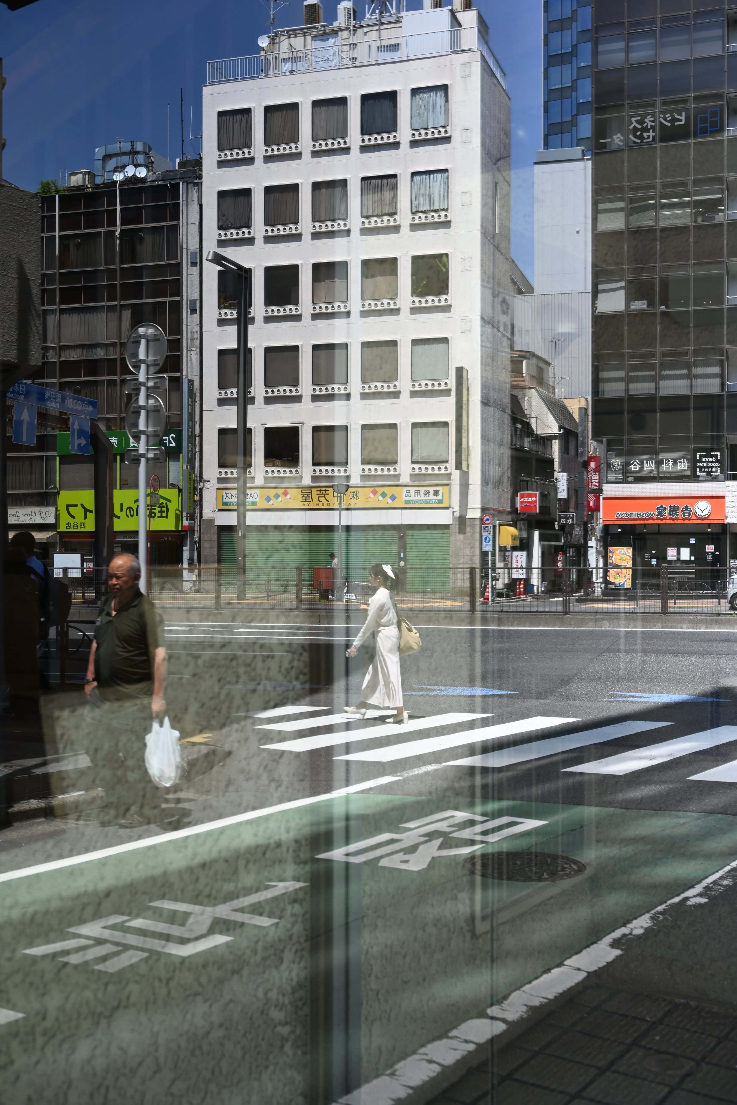 City street scene with people crossing at a crosswalk, buildings in the background displaying signage, and reflections on a glass window.