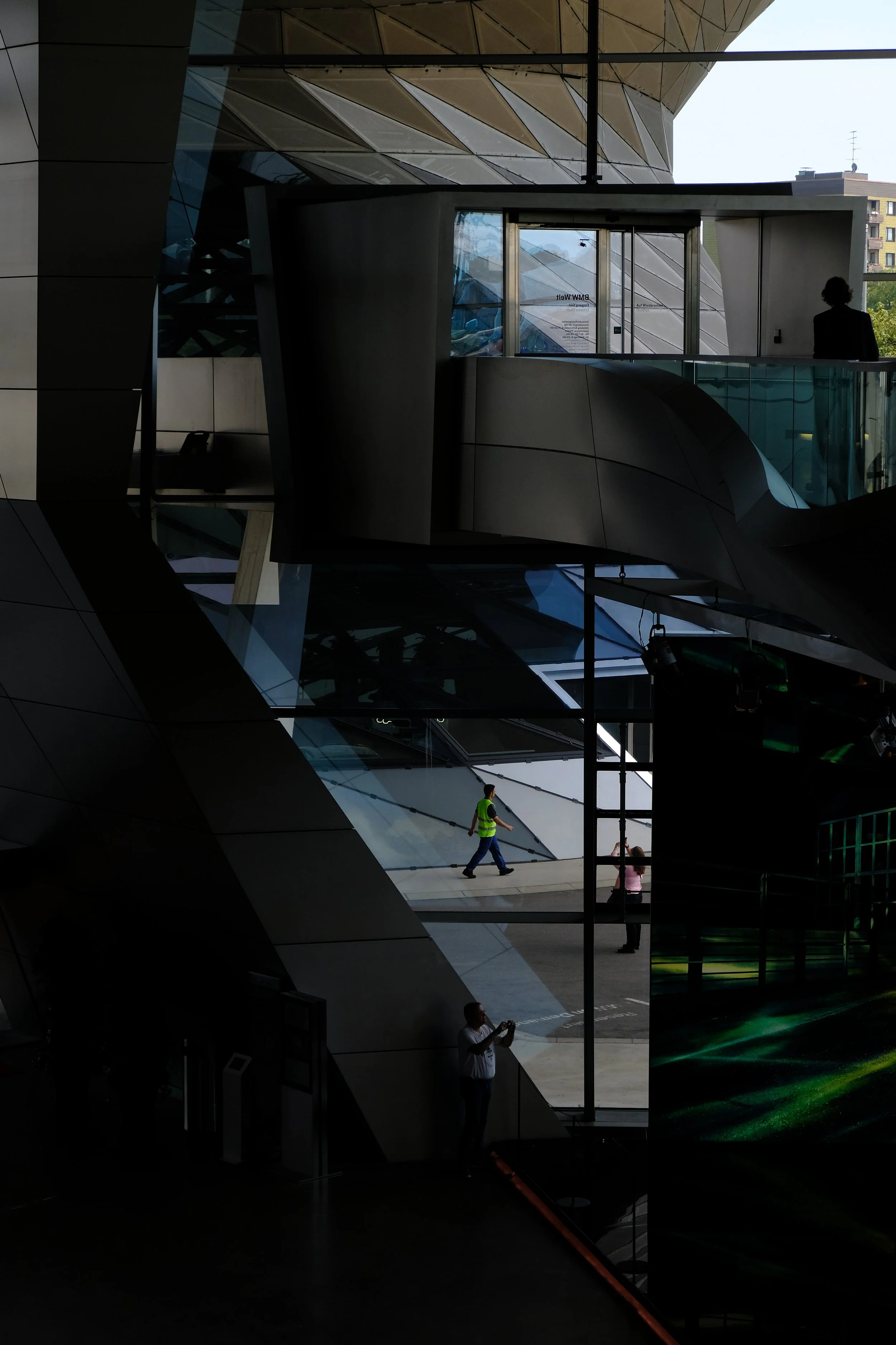 Interior view of a modern building with dark, angular architecture and large glass windows. Two people are visible: a woman on the upper level and a man on the lower level, both inside the building, with one taking a photo.