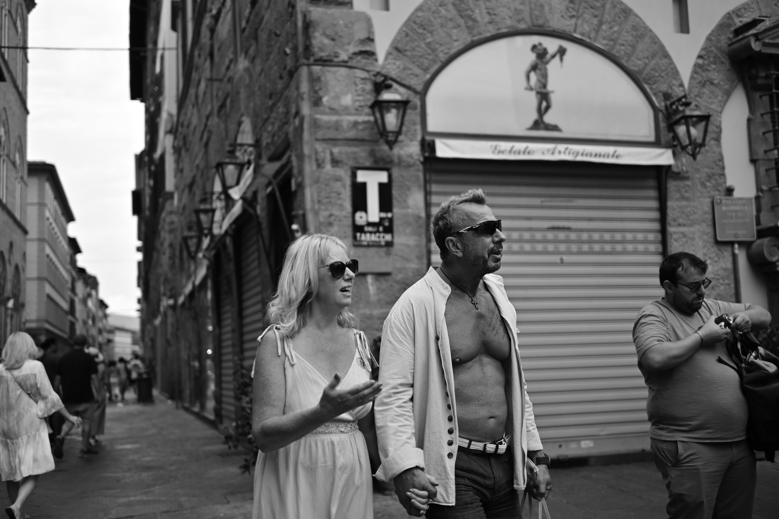 A black and white street scene with three people walking, two men and a woman, holding hands. The woman is talking, wearing sunglasses and a light dress. One man is shirtless with sunglasses and a light jacket, the other man is looking at his camera or phone. In the background, there are buildings and shops with Roll-up shutters.
