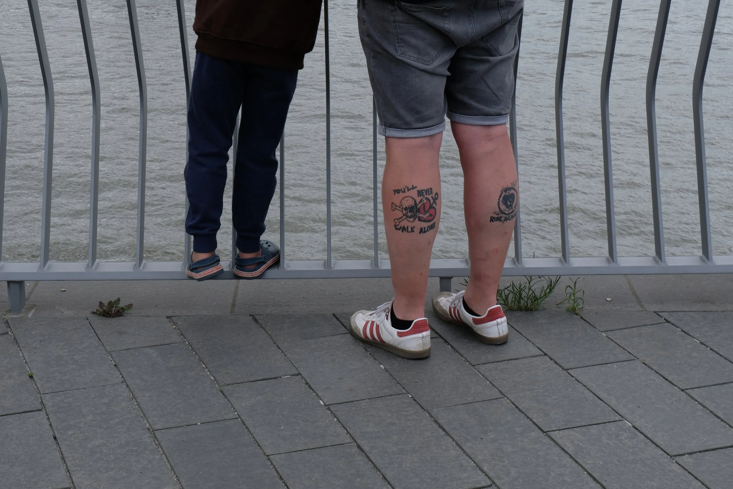 Two people standing by a railing overlooking a body of water. One person is a child with dark pants and sneakers, the other is an adult with tattoos on their legs, wearing shorts and sneakers.