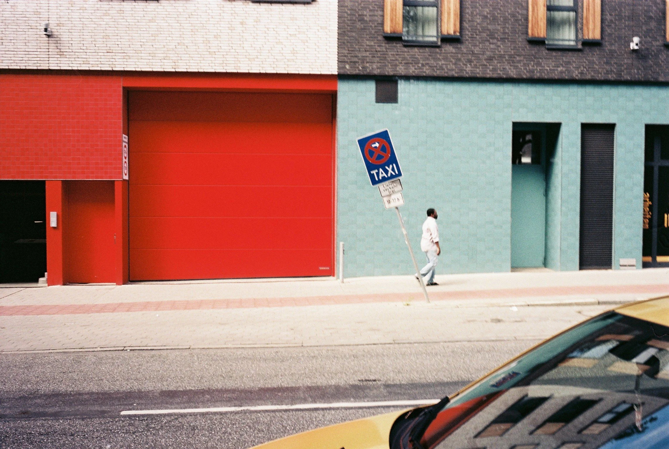 Person walking on sidewalk beside a parked yellow taxi, with a building featuring red, teal, and dark brown sections, and a tilted no-parking sign.