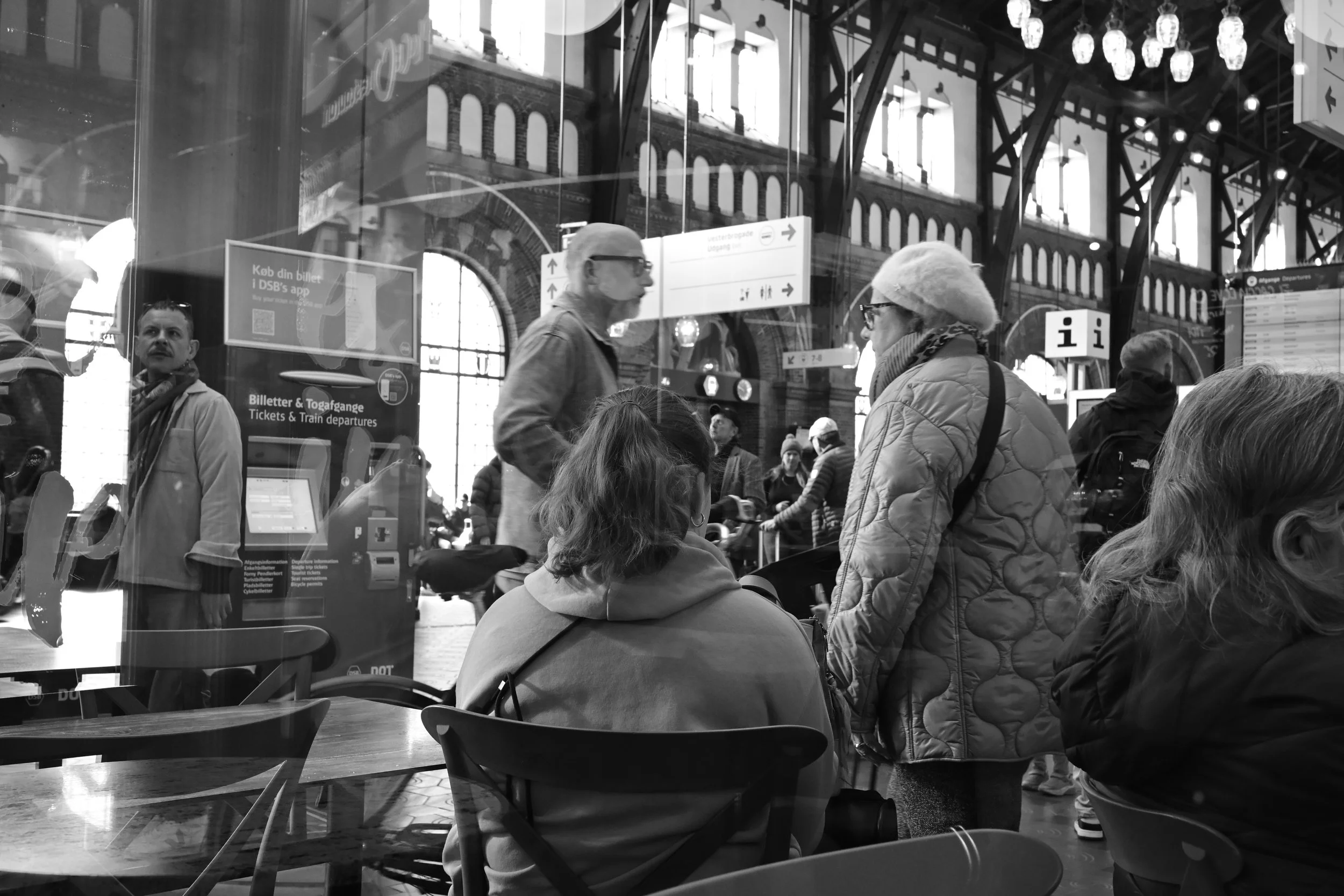 People waiting at a train station, viewed through a glass window, with some sitting and some standing, and large station architecture visible in the background.