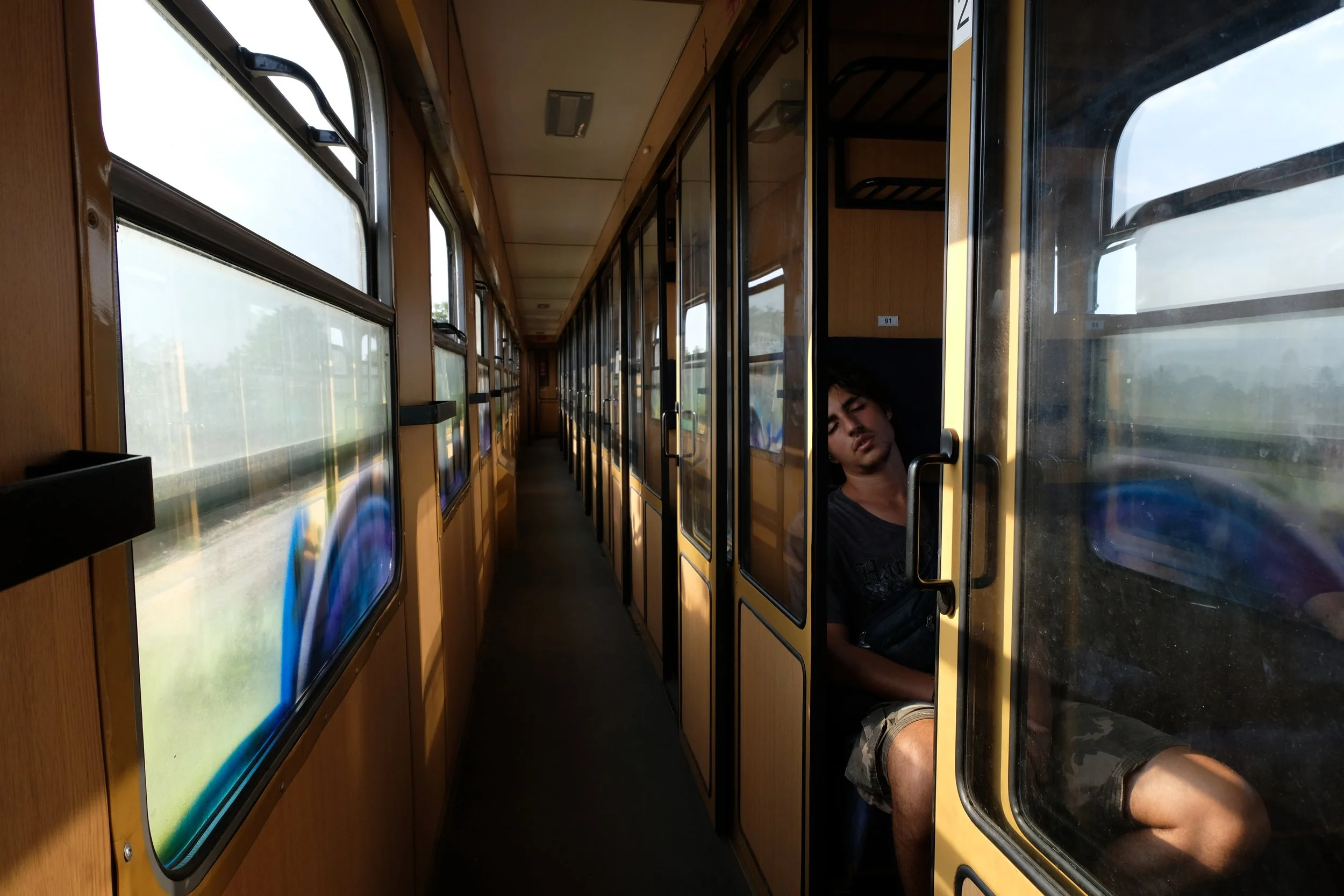 A young man sitting on a train, resting with eyes closed, next to windowed compartment doors on a narrow corridor train interior.