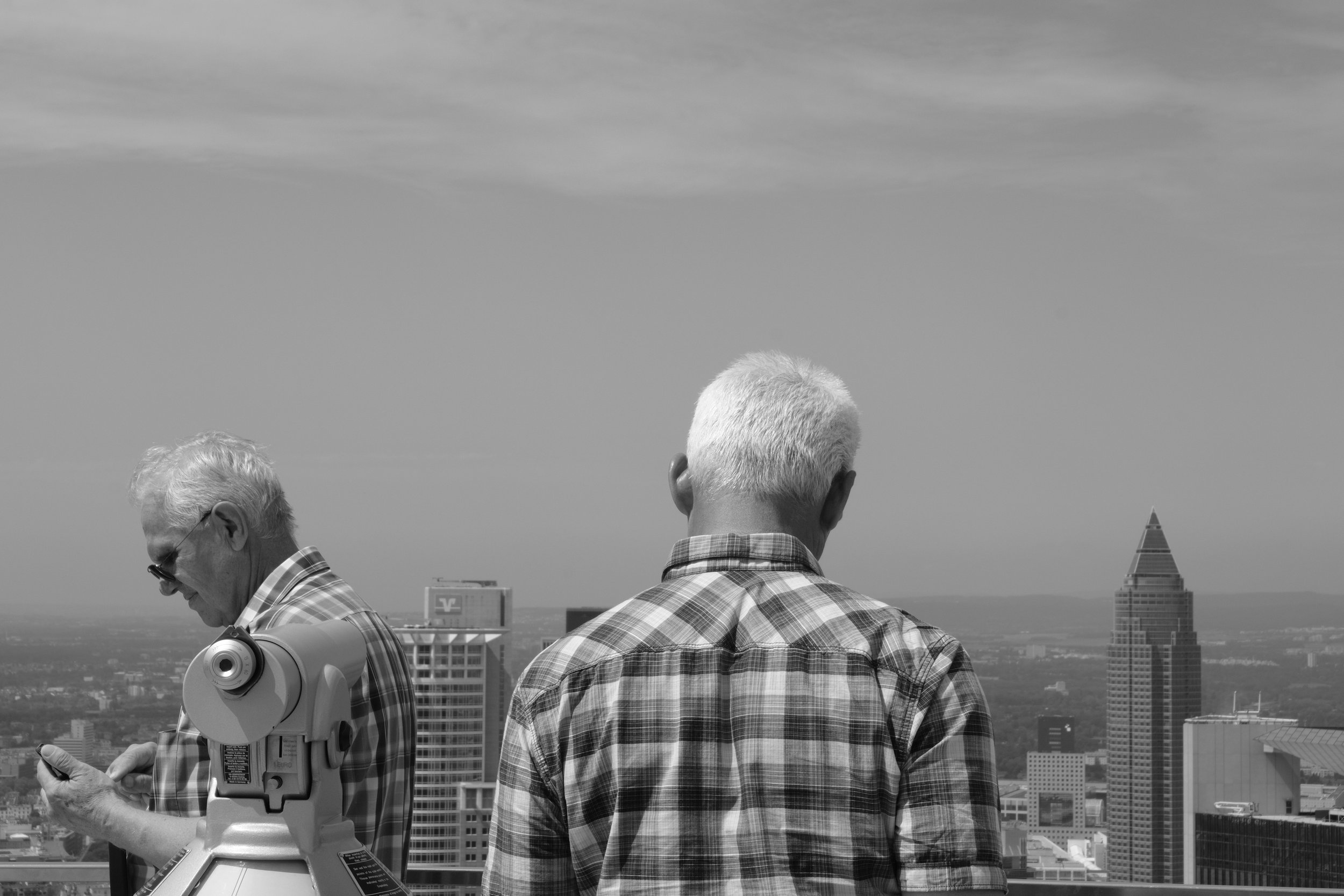 Two elderly men with white hair, wearing plaid shirts, standing on a rooftop with a city skyline in the background. One man is looking at his phone, the other is facing away from the camera. A telescope is also visible in the foreground.