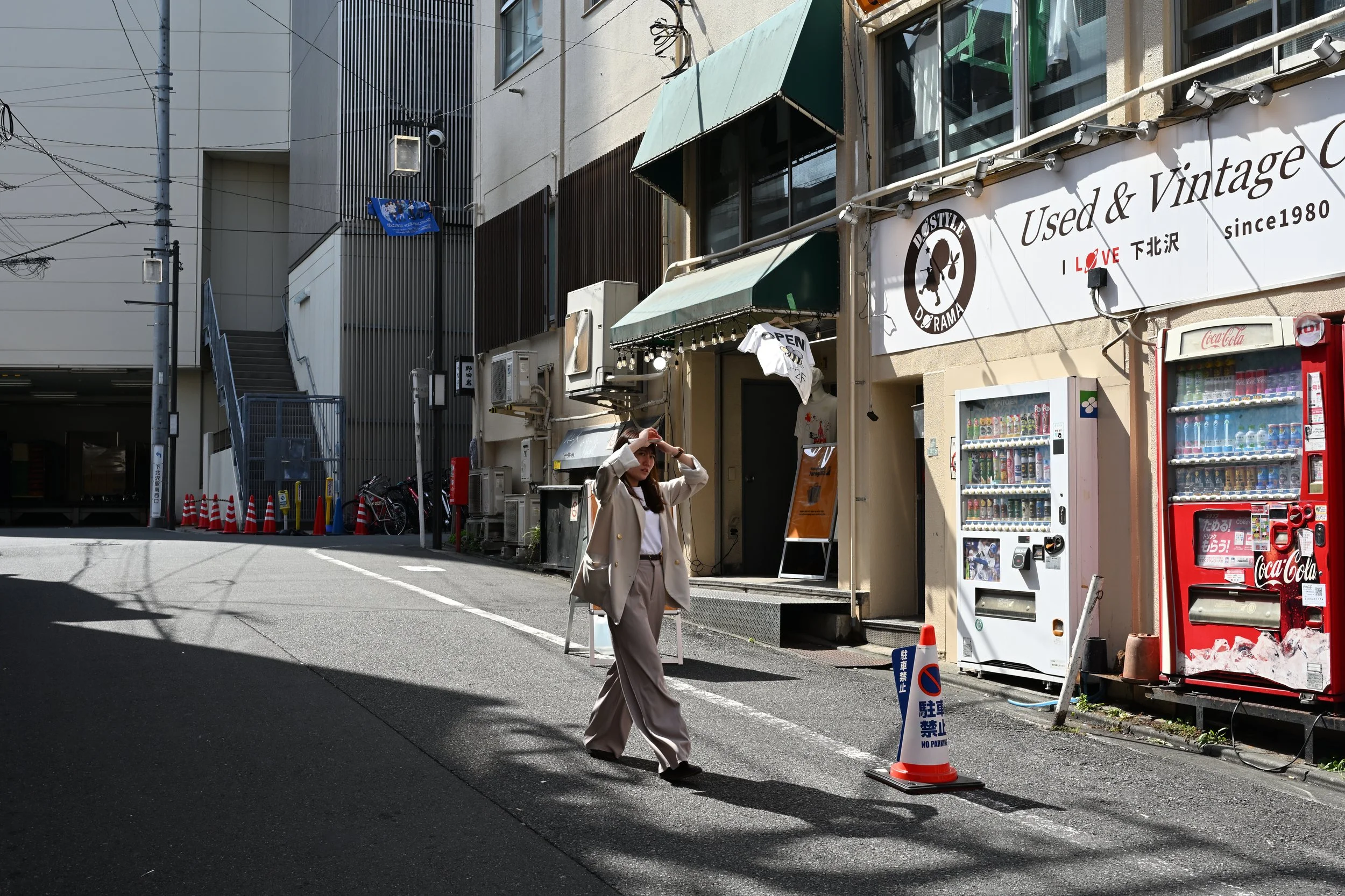 A woman with a beige coat and wide pants walks across a street in an urban area. The street has vending machines and a used and vintage shop with a sign that reads "Used & Vintage C". The background shows buildings, wires, and a staircase.