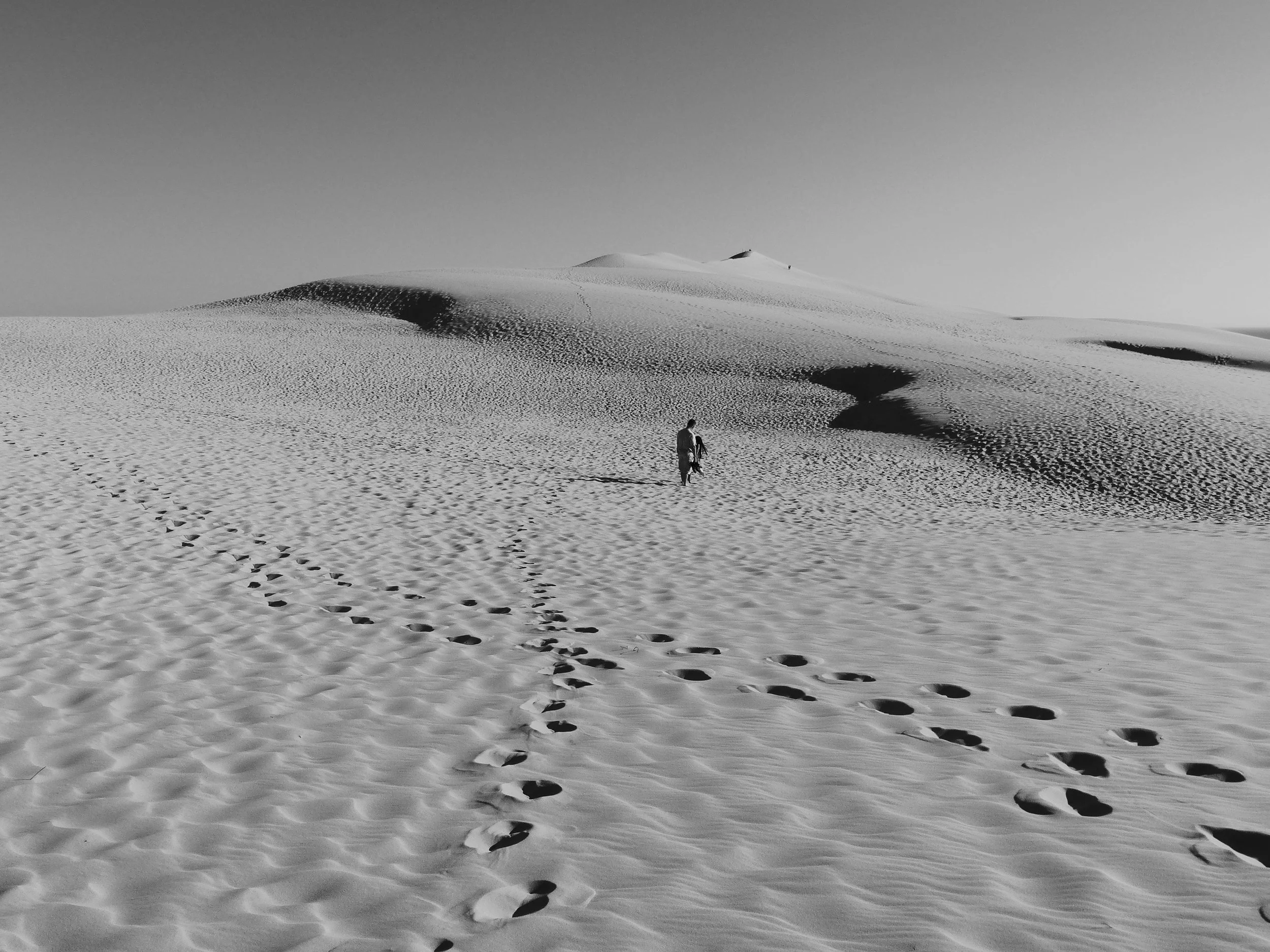 A person walking through a desert with sand dunes, leaving footprints in the sand, under a clear sky.