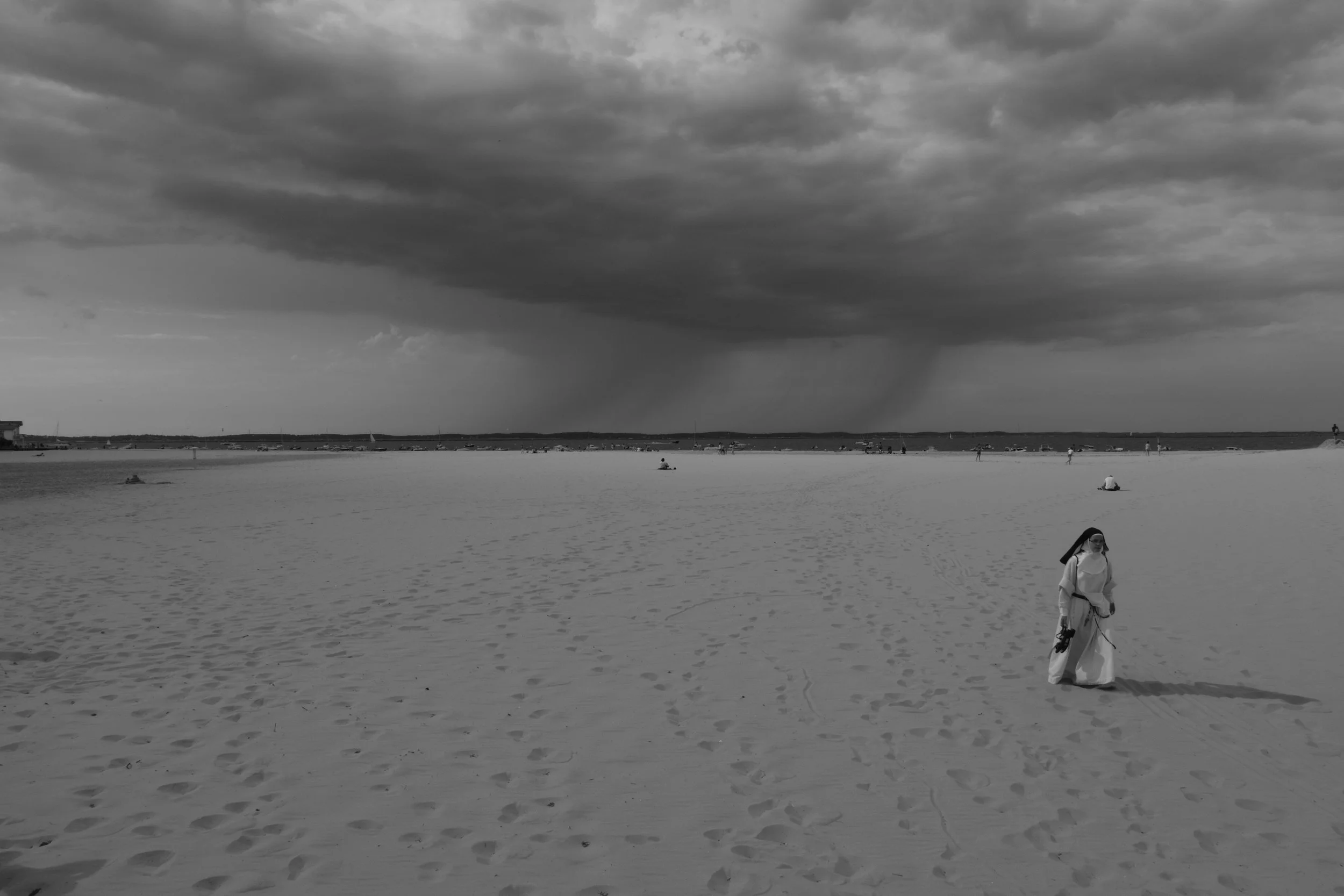 A person dressed in traditional white attire walking on a beach with a cloudy sky and rain in the distance.