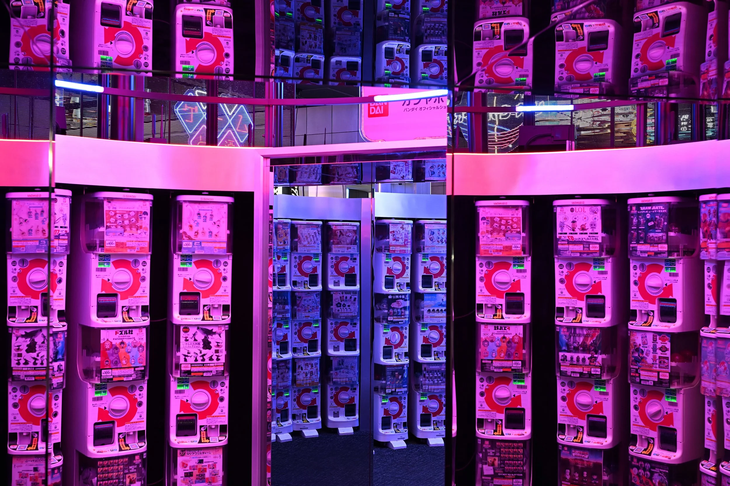 Multiple pink and white capsule toy vending machines in a brightly lit arcade with neon signs, showcasing a vibrant, colorful atmosphere.