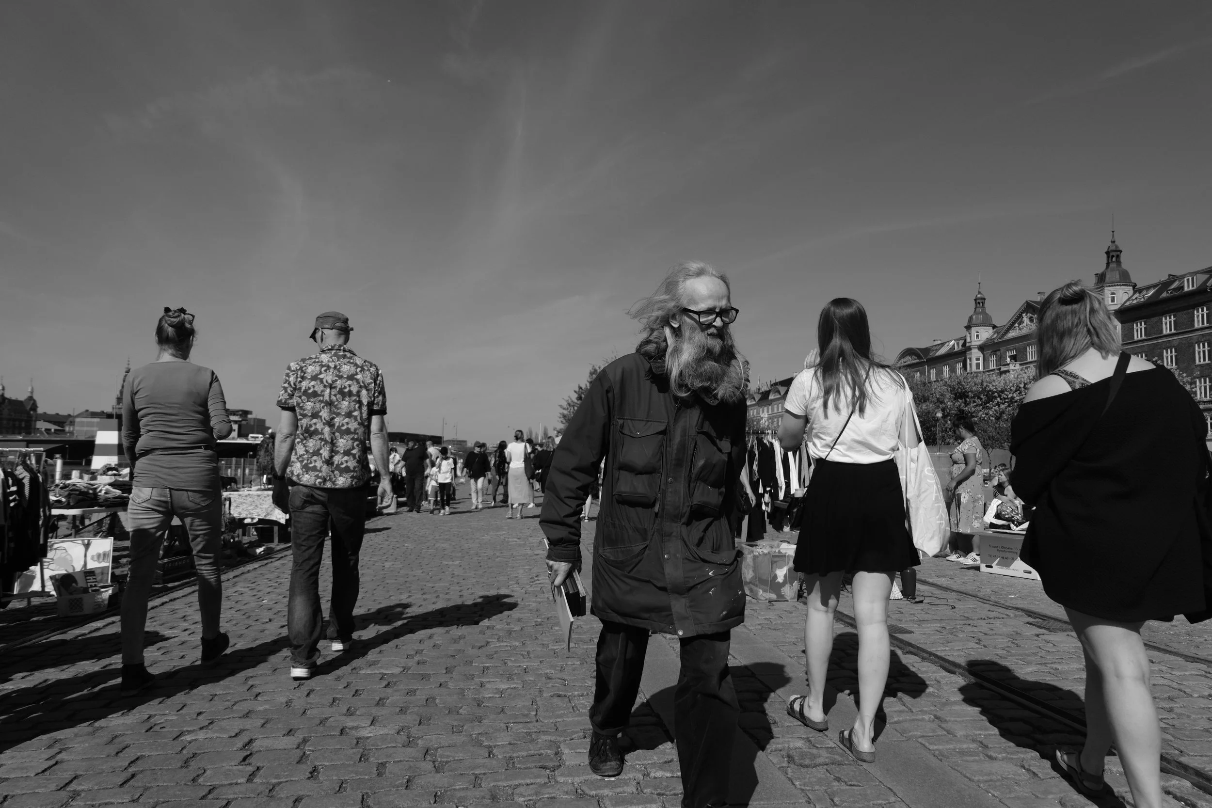 People walking and browsing at an outdoor market on a sunny day in a city, with historic buildings in the background.