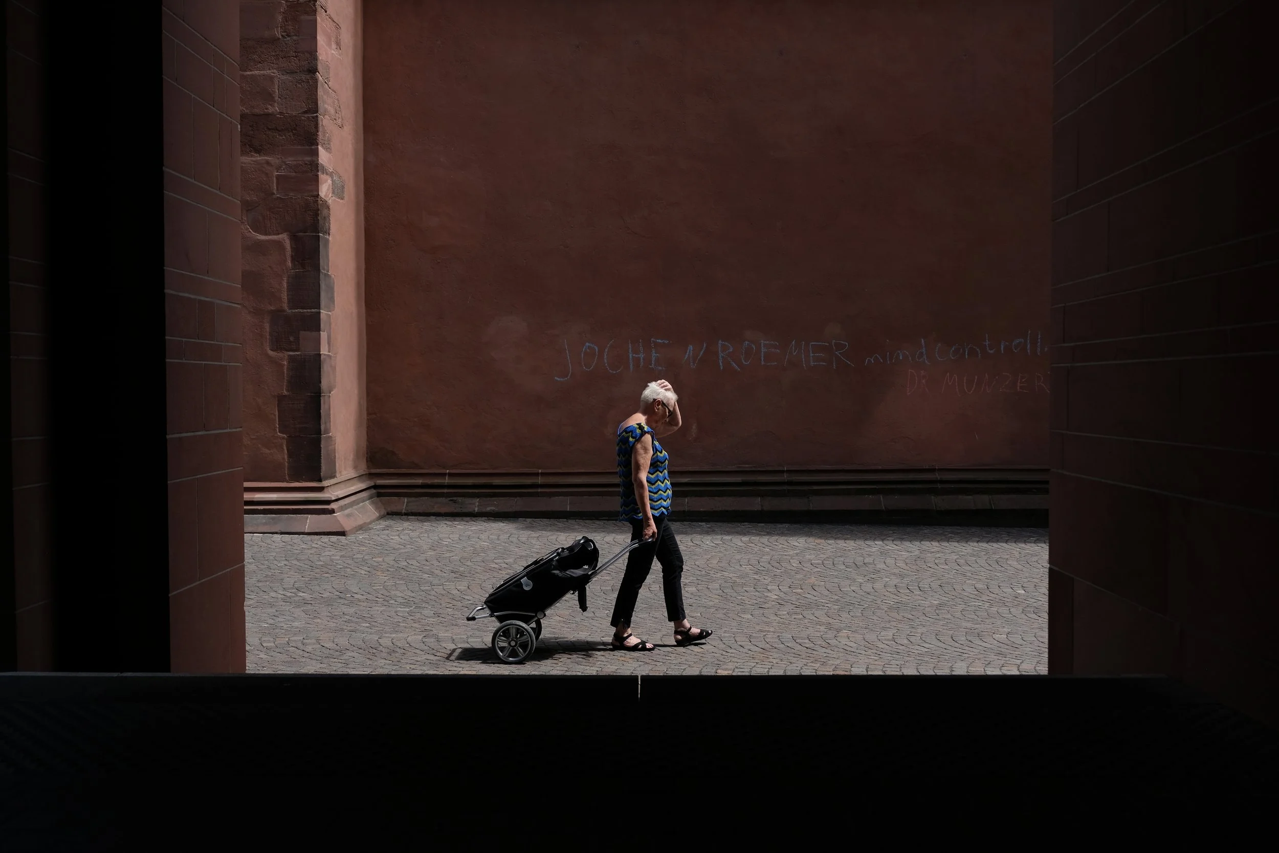 An elderly woman with white hair, wearing a blue patterned sleeveless top, black pants, and sandals, walking through a tunnel while pulling a black rolling suitcase.
