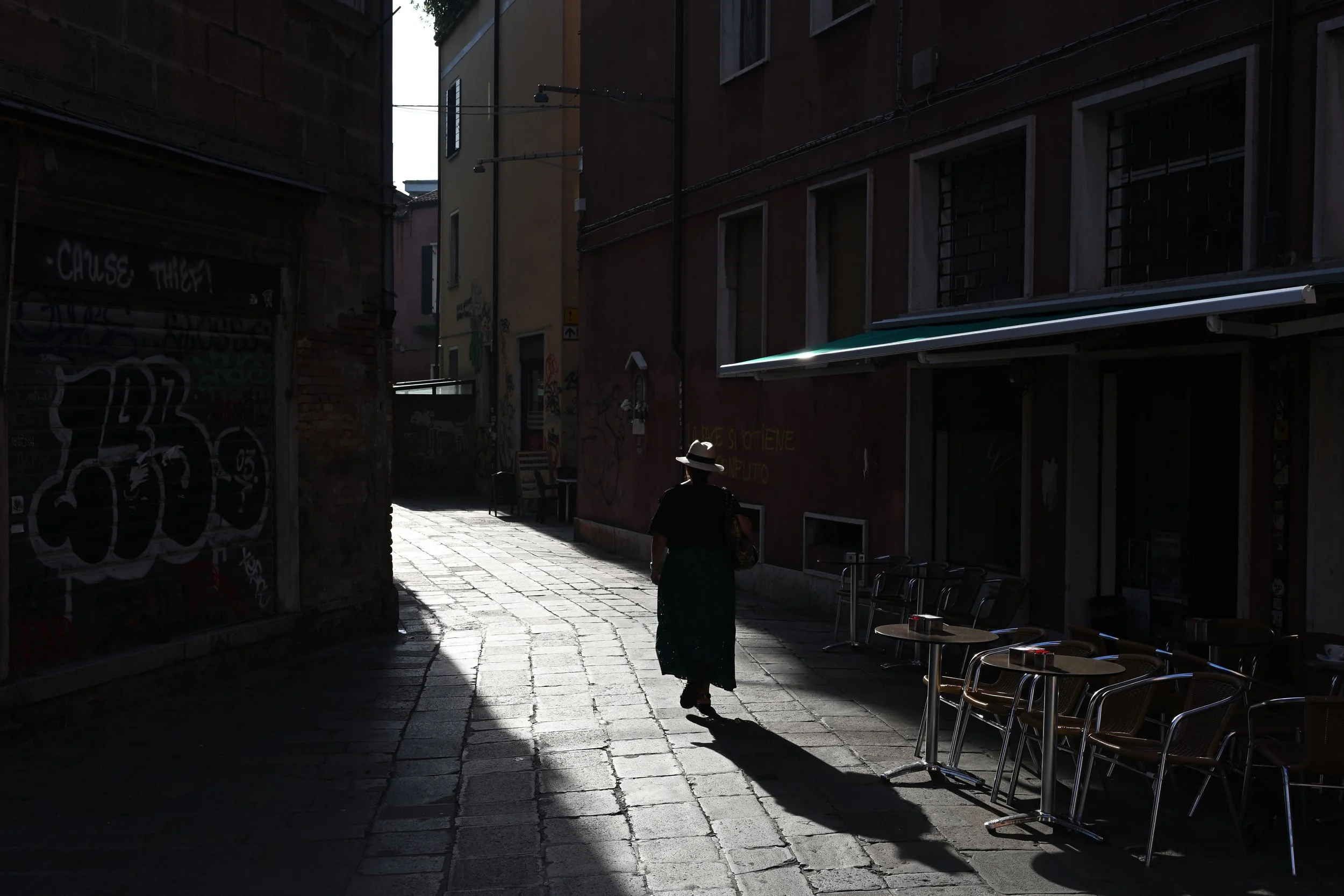 A person walking down a narrow, shadowed alleyway with outdoor chairs stacked on the right side, surrounded by tall buildings with graffiti on the walls.