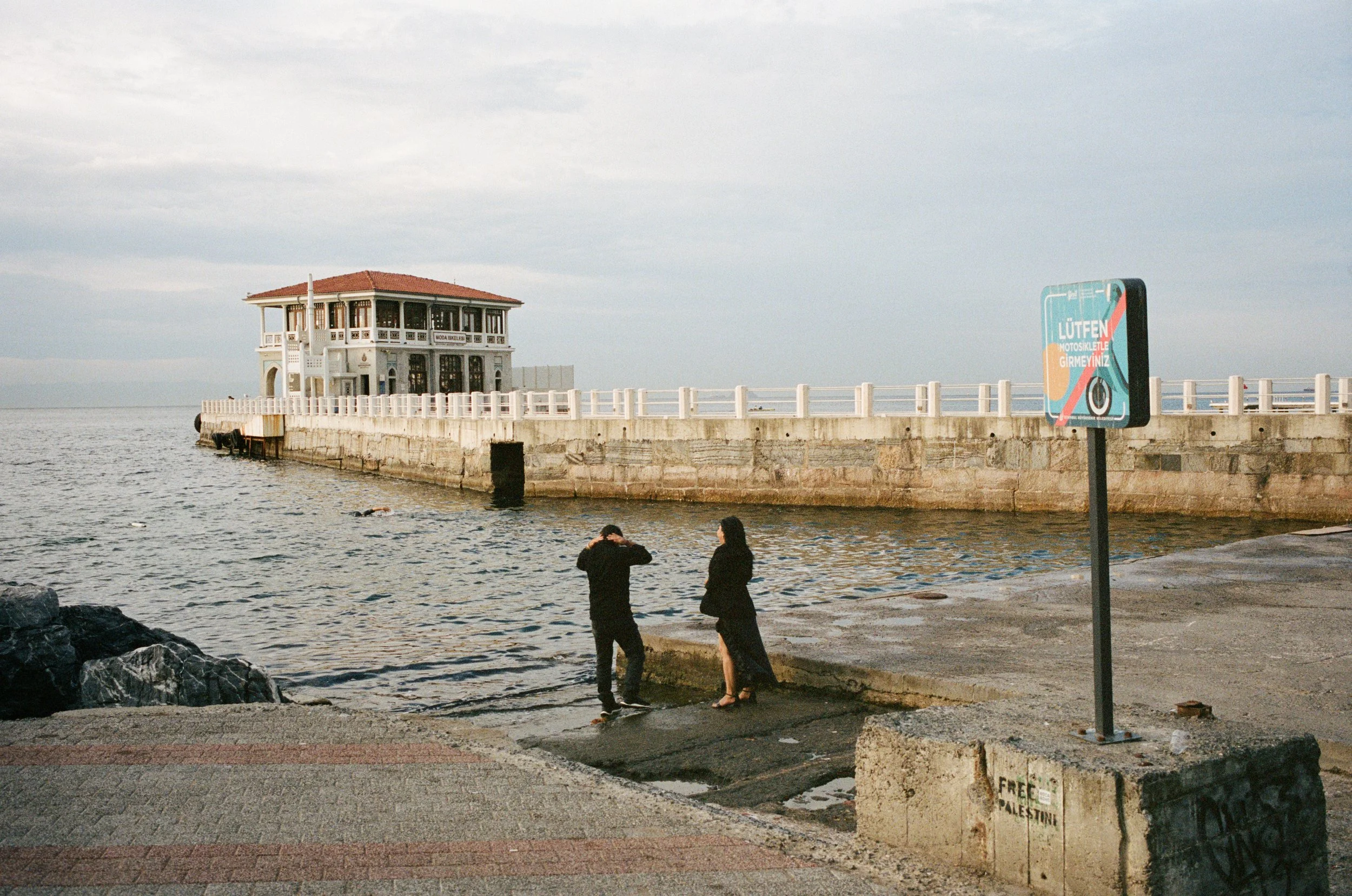 A seaside scene with a stone dock and two people talking, one man and one woman, near the water. In the background, there is a two-story restaurant or house on the pier, and a sign with Turkish writing. The sky is partly cloudy.