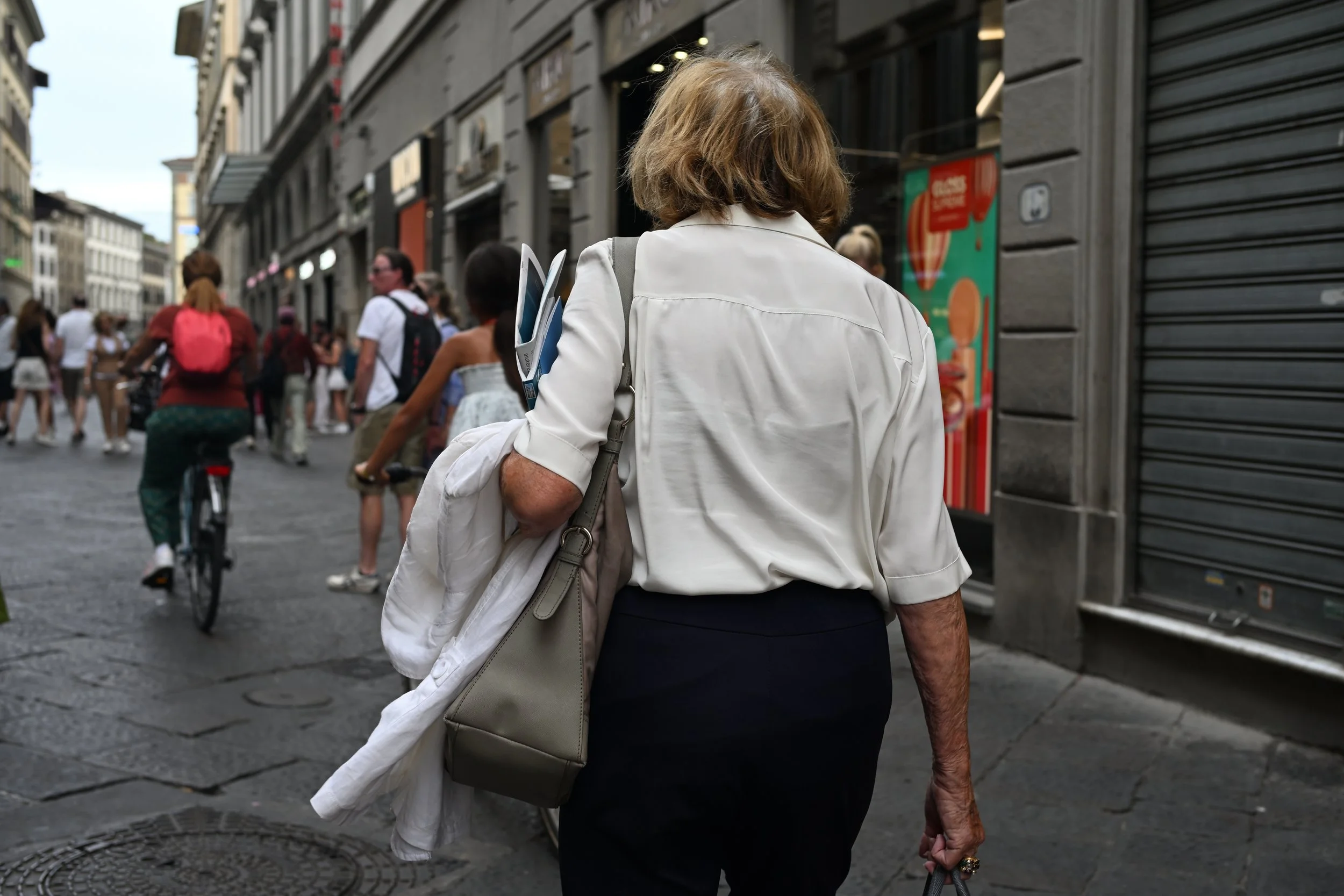 Back view of a woman with short light brown hair wearing a white blouse and black skirt walking on a busy city street with people, shops, and buildings.