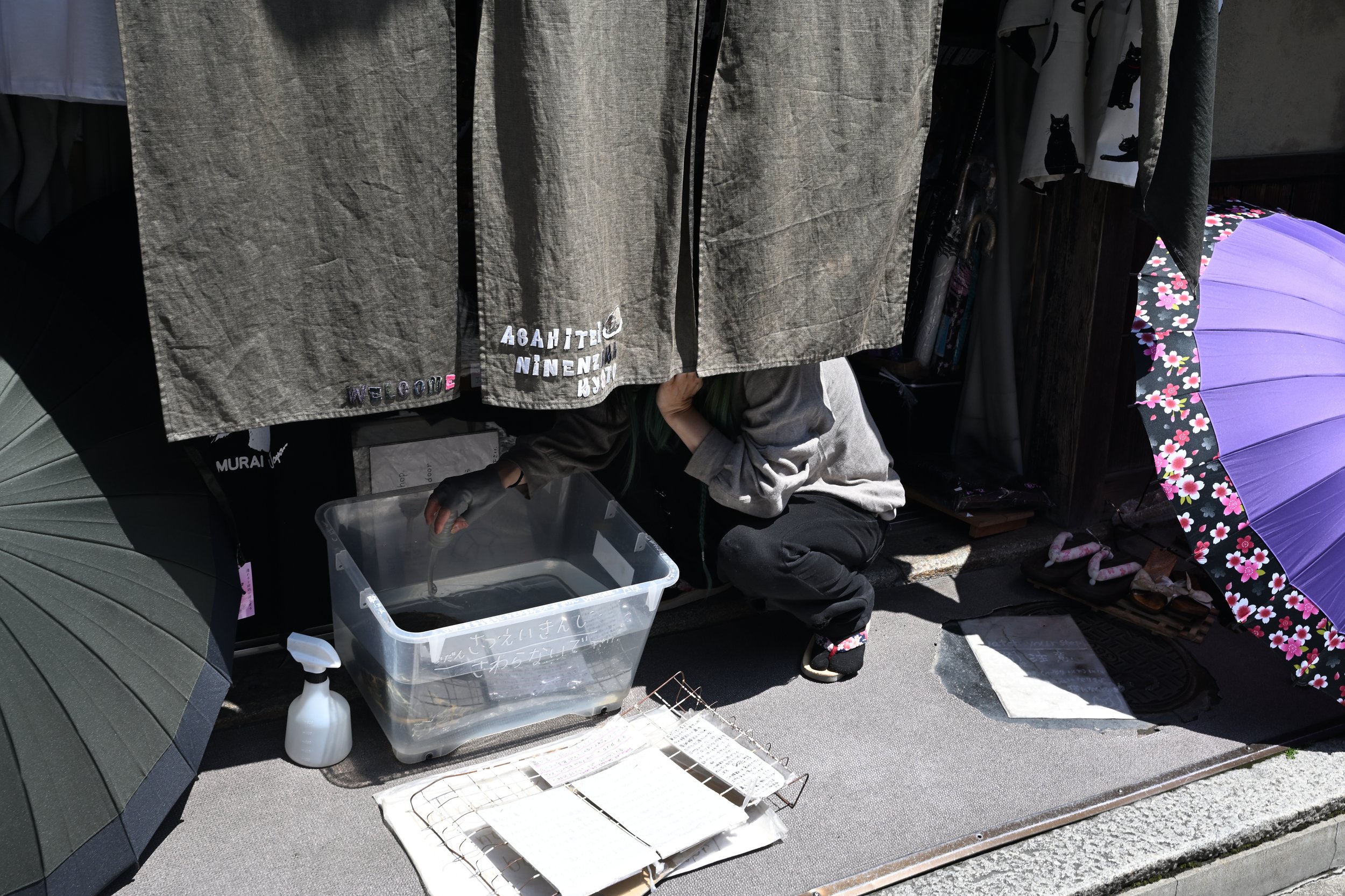 A person crouched under a makeshift shelter with hanging fabric and umbrellas, next to a plastic container with a Japanese sign, and a spray bottle, on a sidewalk.