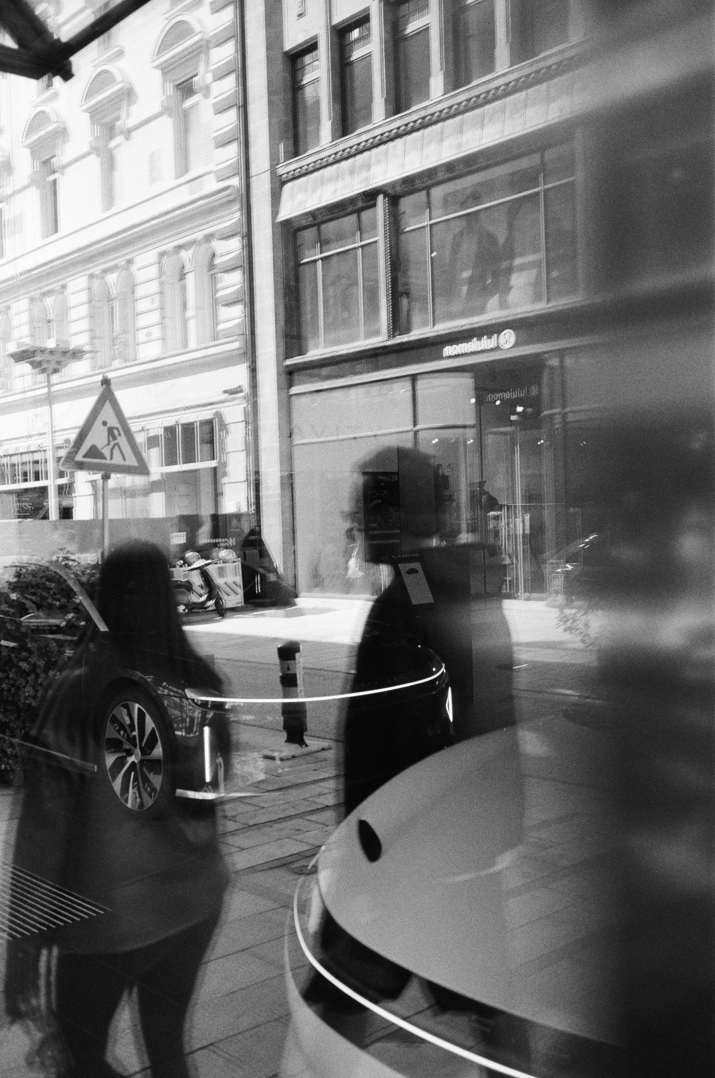 Reflection of two people in a store window, with buildings, parked cars, and a street scene outside in the background, in black and white.