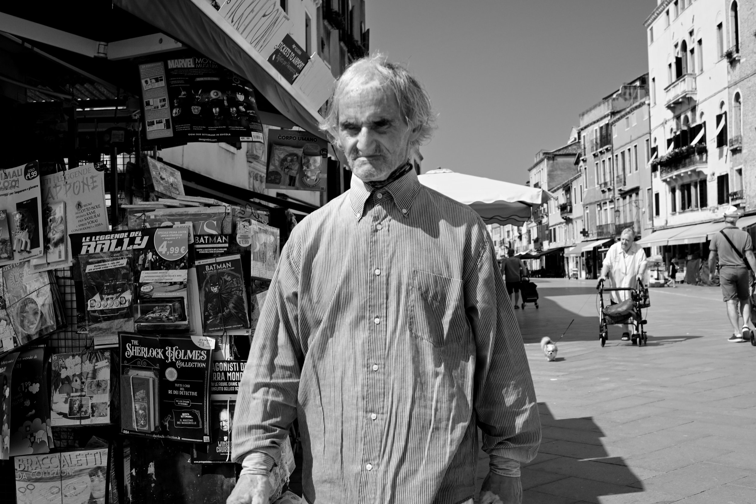 A black and white photo of an elderly man with light-colored hair and a beard, standing on a busy city street near a kiosk displaying comic books and magazines. The man wears a striped button-up shirt and looks directly at the camera with a serious expression. In the background, there are other pedestrians, some of whom are pushing strollers, walking along the sidewalk.