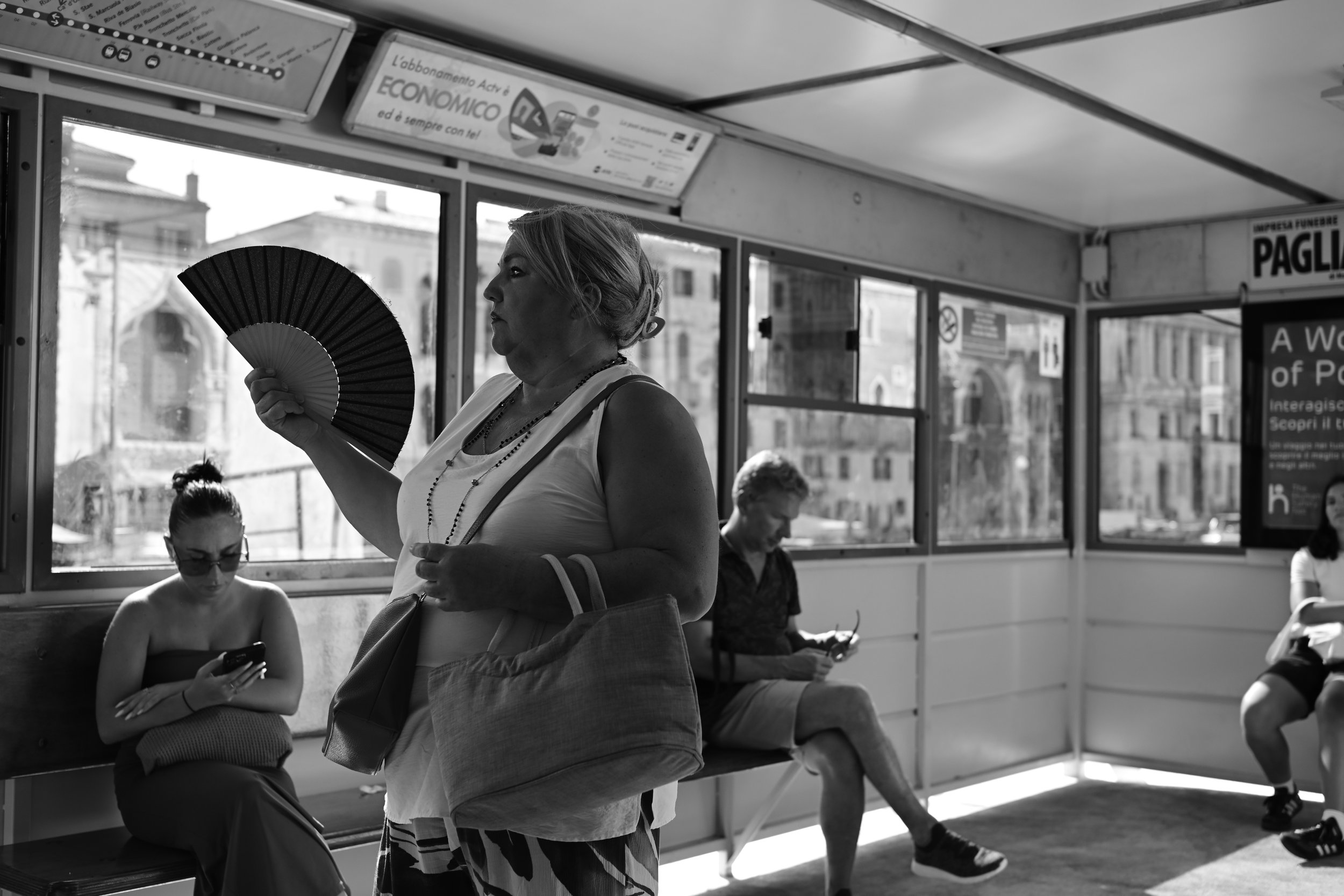 A woman standing inside a bus holding a fan, with three people seated near her, two women and one man, all looking at their phones or sunglasses, with city buildings visible through the bus windows in the background.