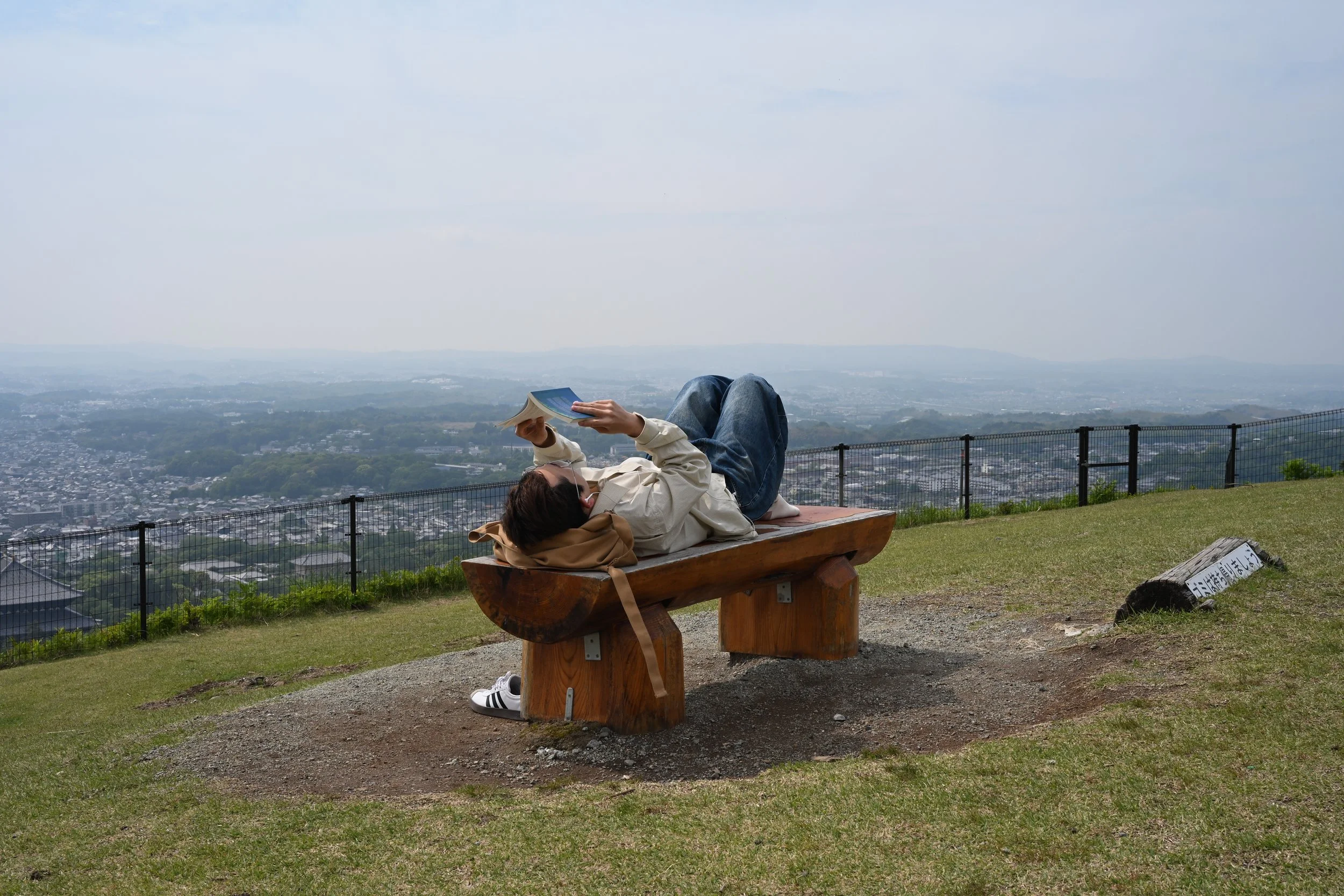 Person lying on a wooden bench, reading a book, overlooking a cityscape and distant hills.