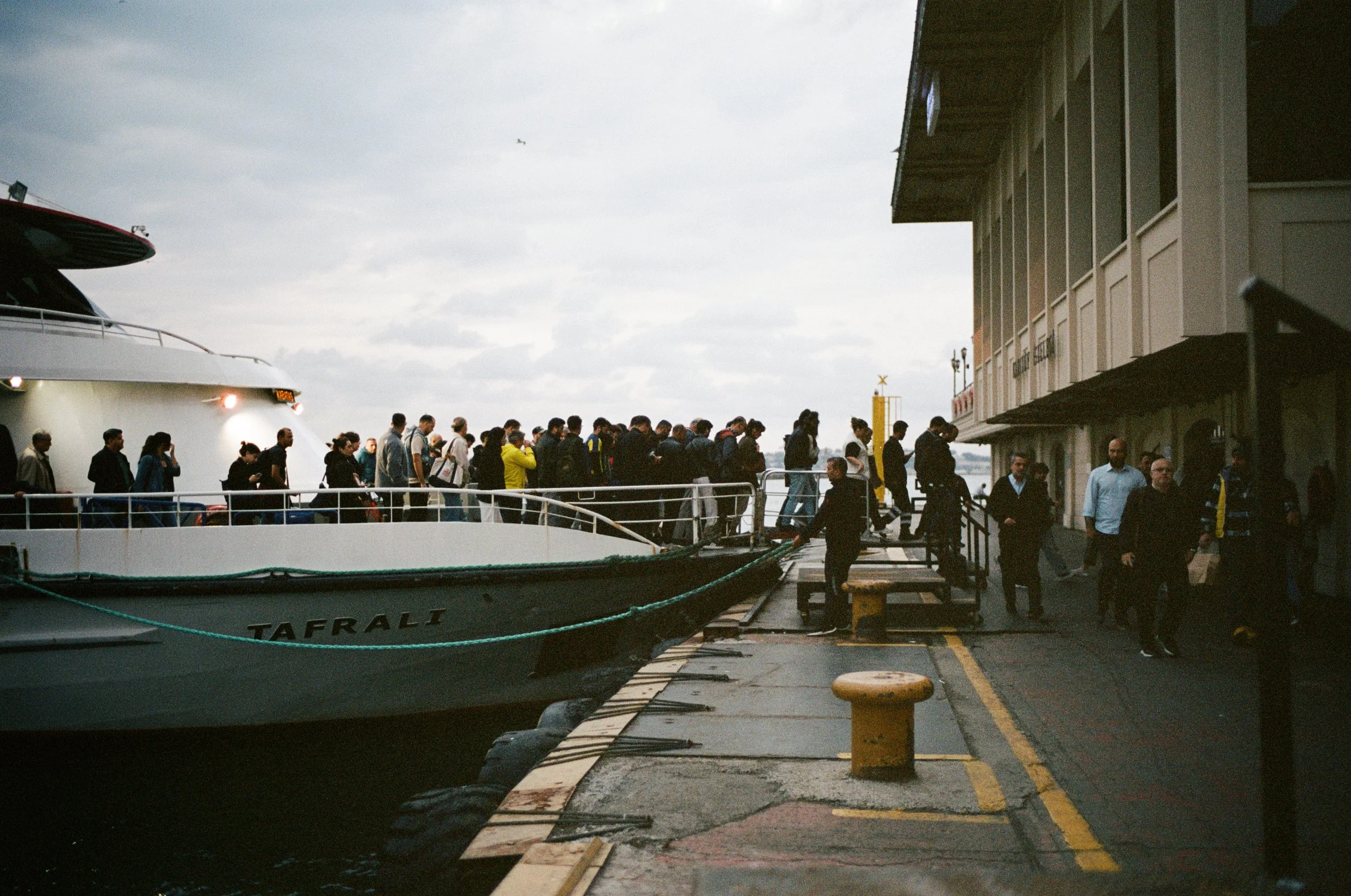 People standing on a dock next to a large yacht named TAFLALI, preparing to board or disembark during cloudy weather.