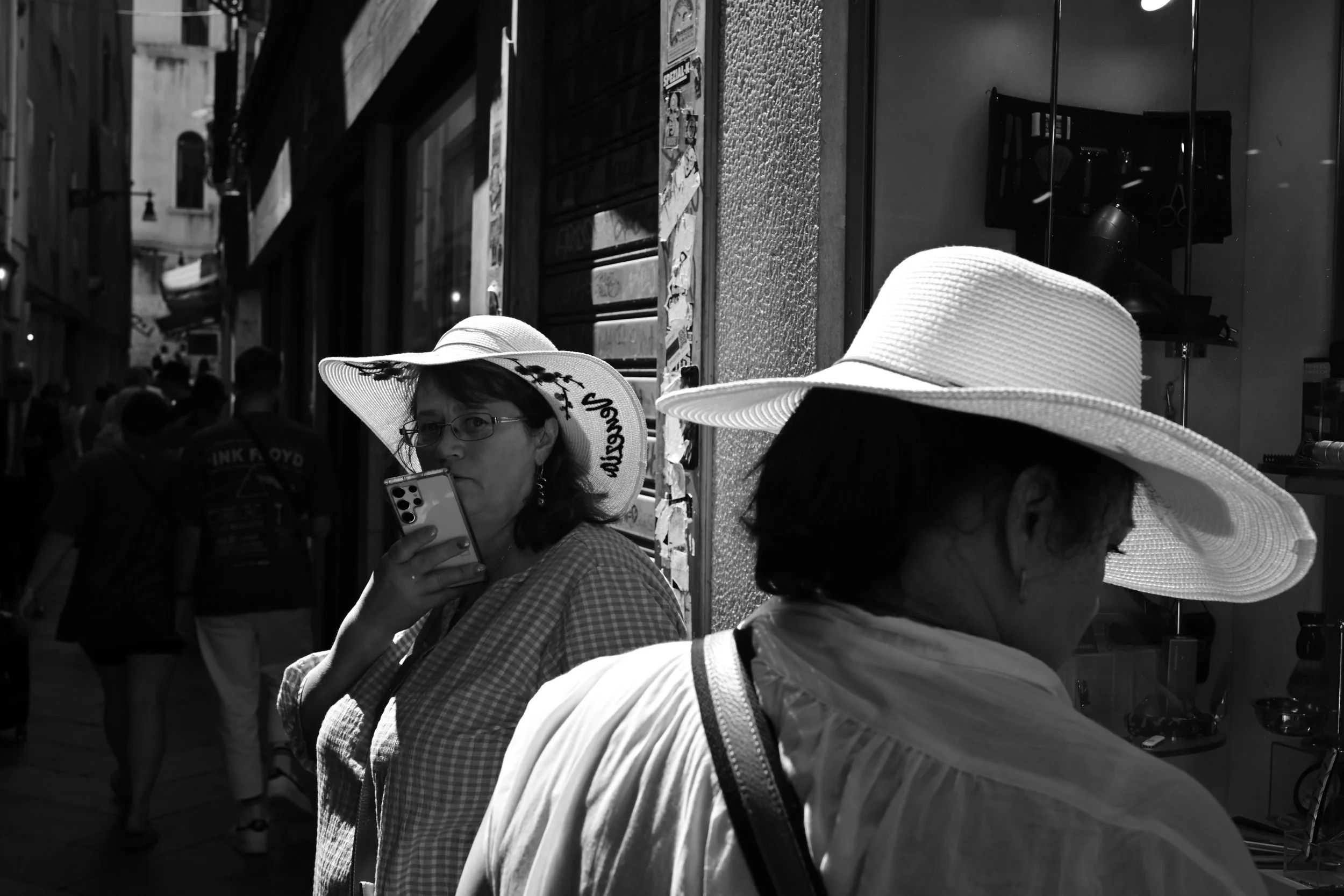 Two women wearing wide-brimmed hats on a busy street, one woman is using a smartphone. The background shows a crowd and storefronts.