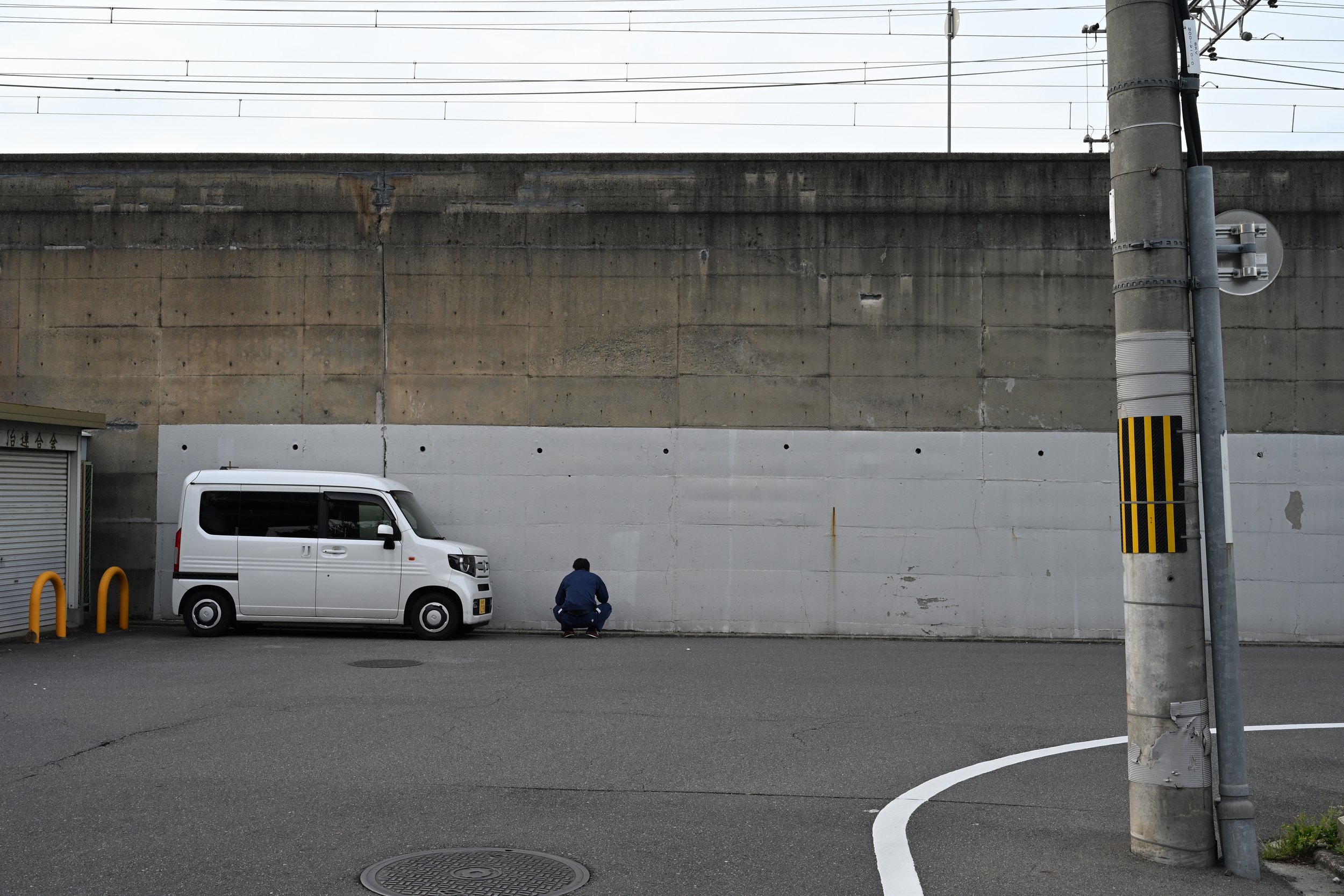 A person squatting next to a white van parked on the side of a street under an overpass wall. There is a utility pole on the right and a wall behind the person and van.