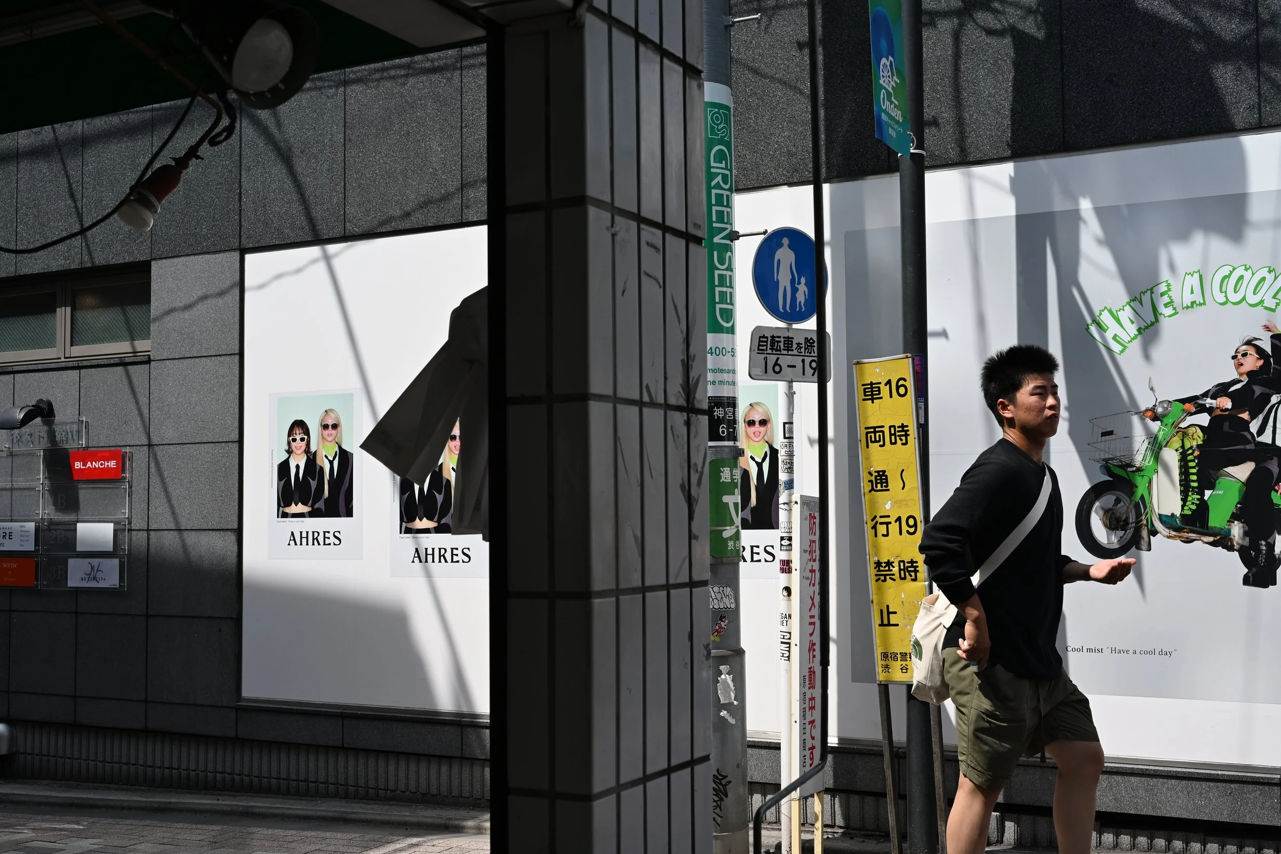 A pedestrian walking past a billboard with cartoon images of three women in sunglasses, one with blonde hair. There are various street signs and advertisements in the background, including a green sign with text, a blue sign indicating a pedestrian crossing, and a yellow sign with Asian characters. The photo is taken in an urban area with a building facade made of gray tiles.