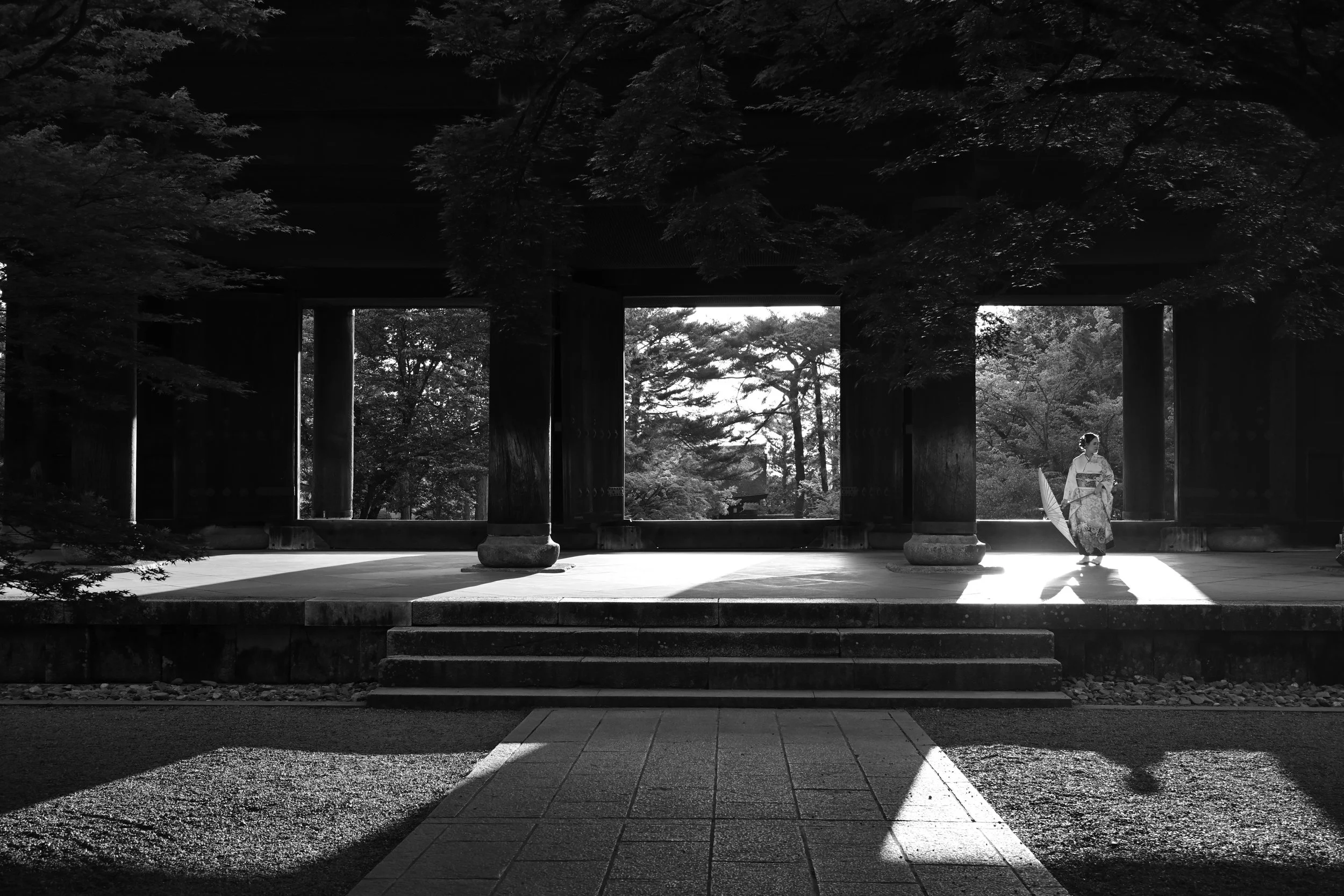 A woman wearing a kimono holds an umbrella and stands on a stage with steps, framed by traditional architecture, with trees visible in the background and sunlight casting shadows.