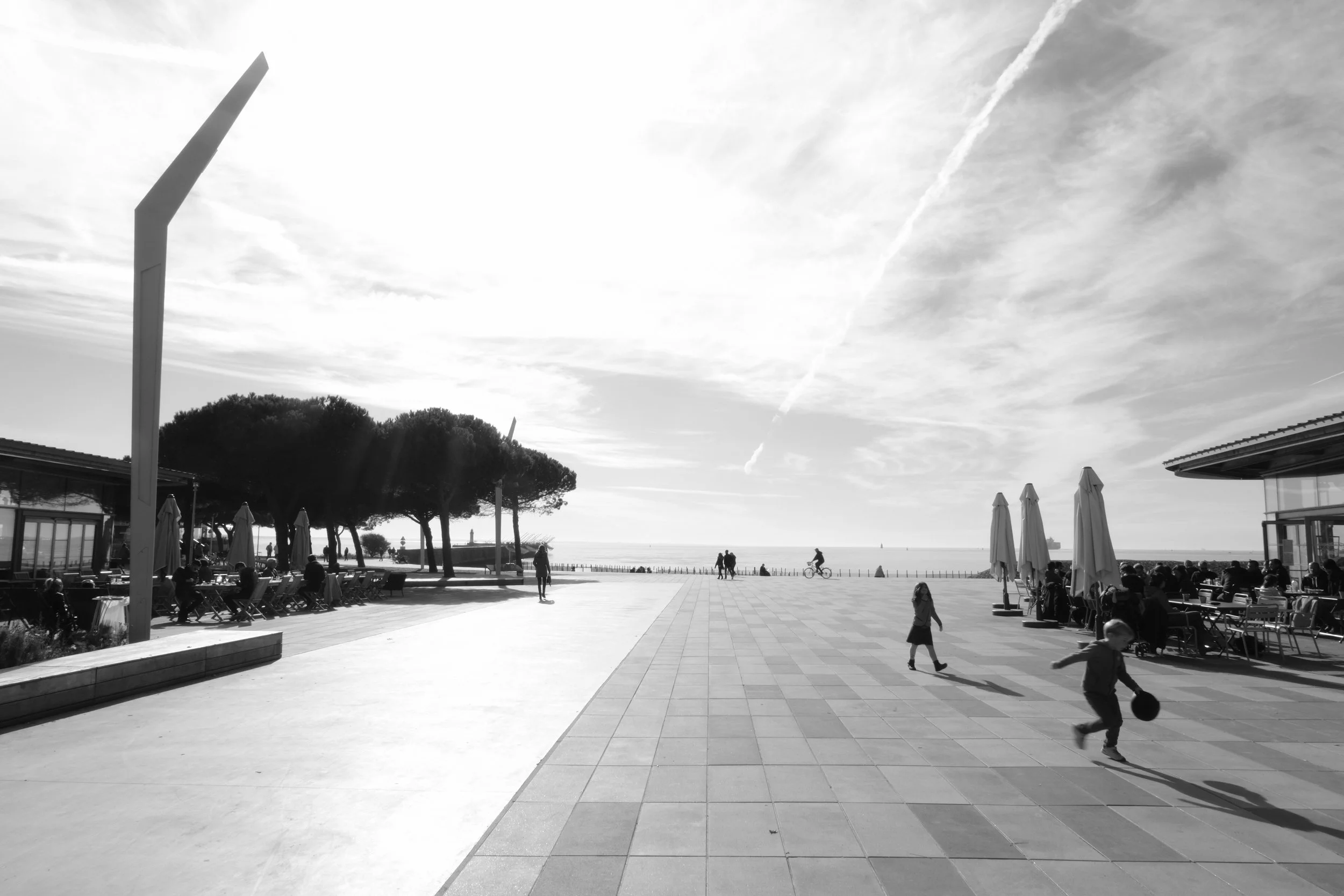 A seaside promenade with people walking, children playing, trees, umbrellas, and a view of the ocean in the background under a partly cloudy sky.