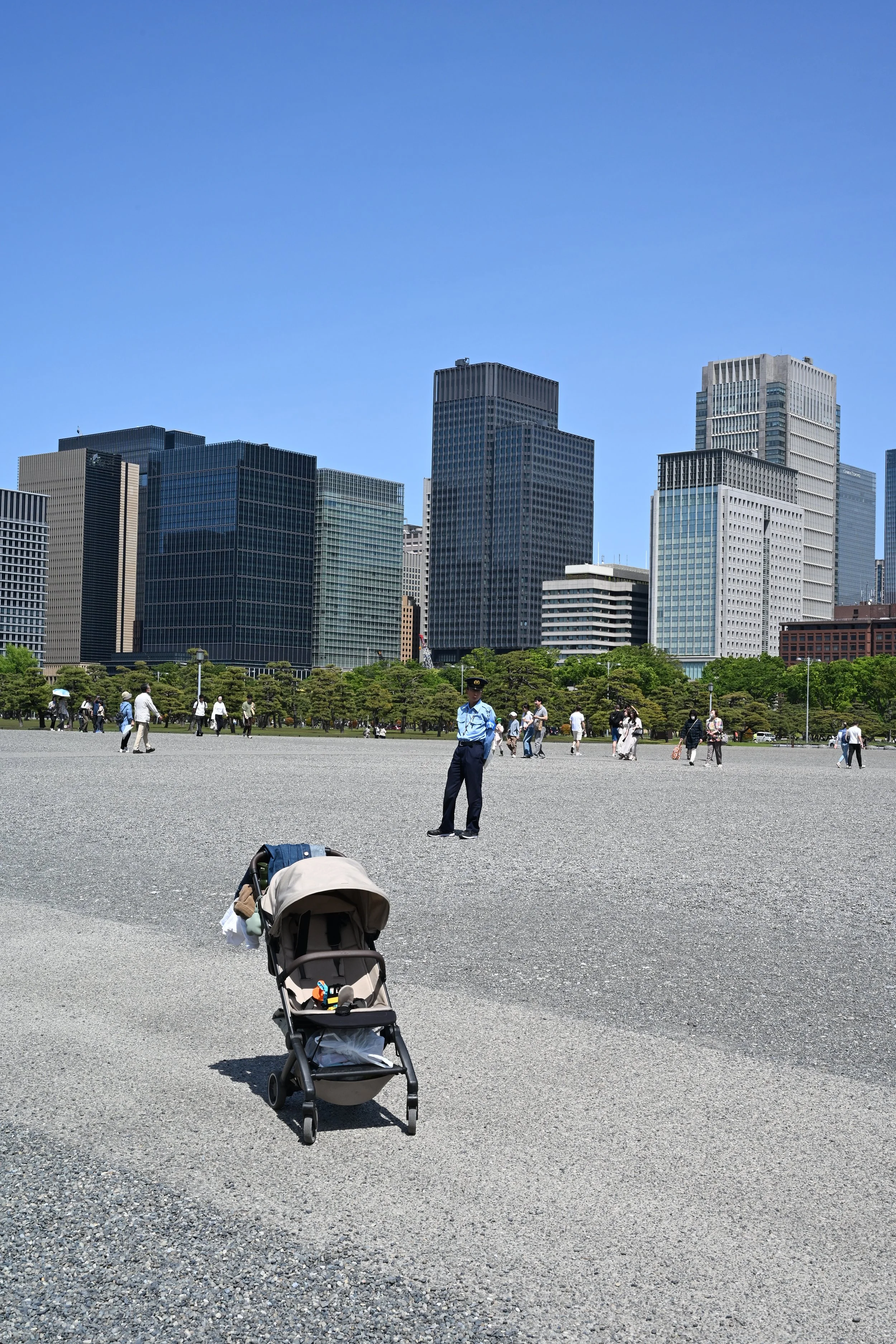 A stroller with a blanket and small toys rests on gravel ground in a park, with people walking and a police officer standing nearby, against a backdrop of tall skyscrapers under a clear blue sky.