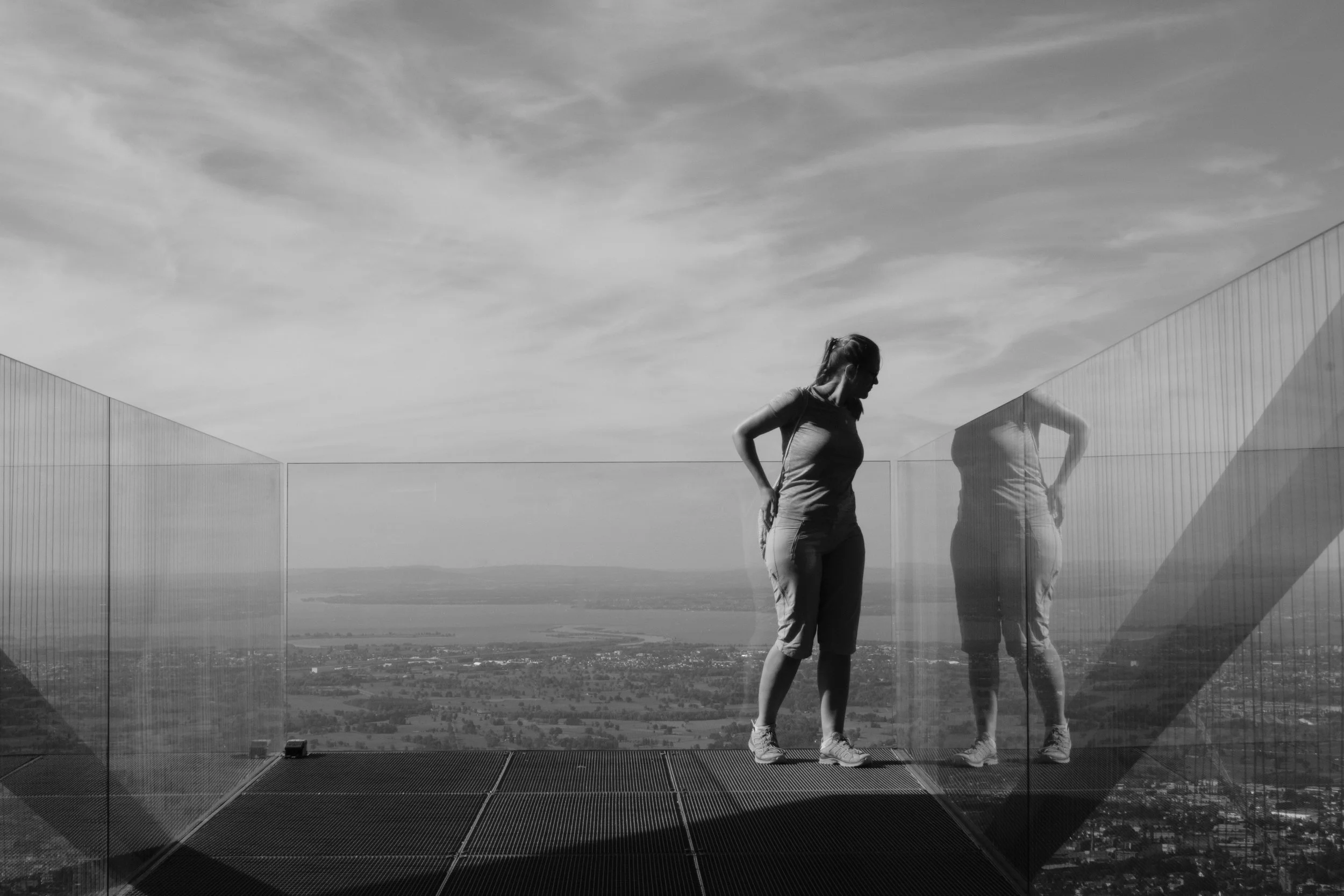 A woman on a viewing platform with glass railings, looking at the scenery below, her reflection visible in the glass.