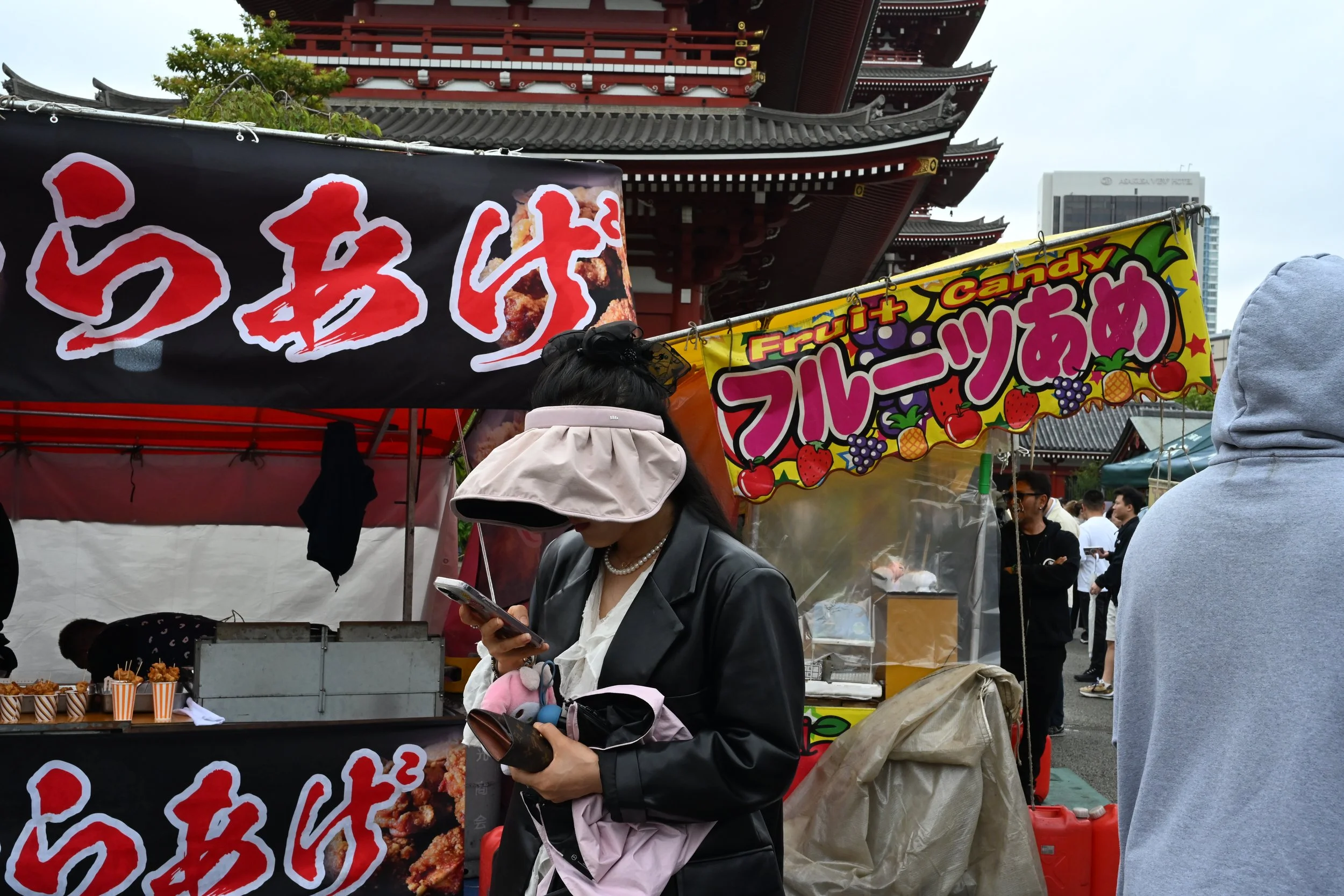 A woman in a leather jacket and wide-brimmed hat stands looking at her phone in front of a food stall at a Japanese festival, with a temple in the background. The stall has colorful signs in Japanese and English, advertising fried chicken and various fruit candies.
