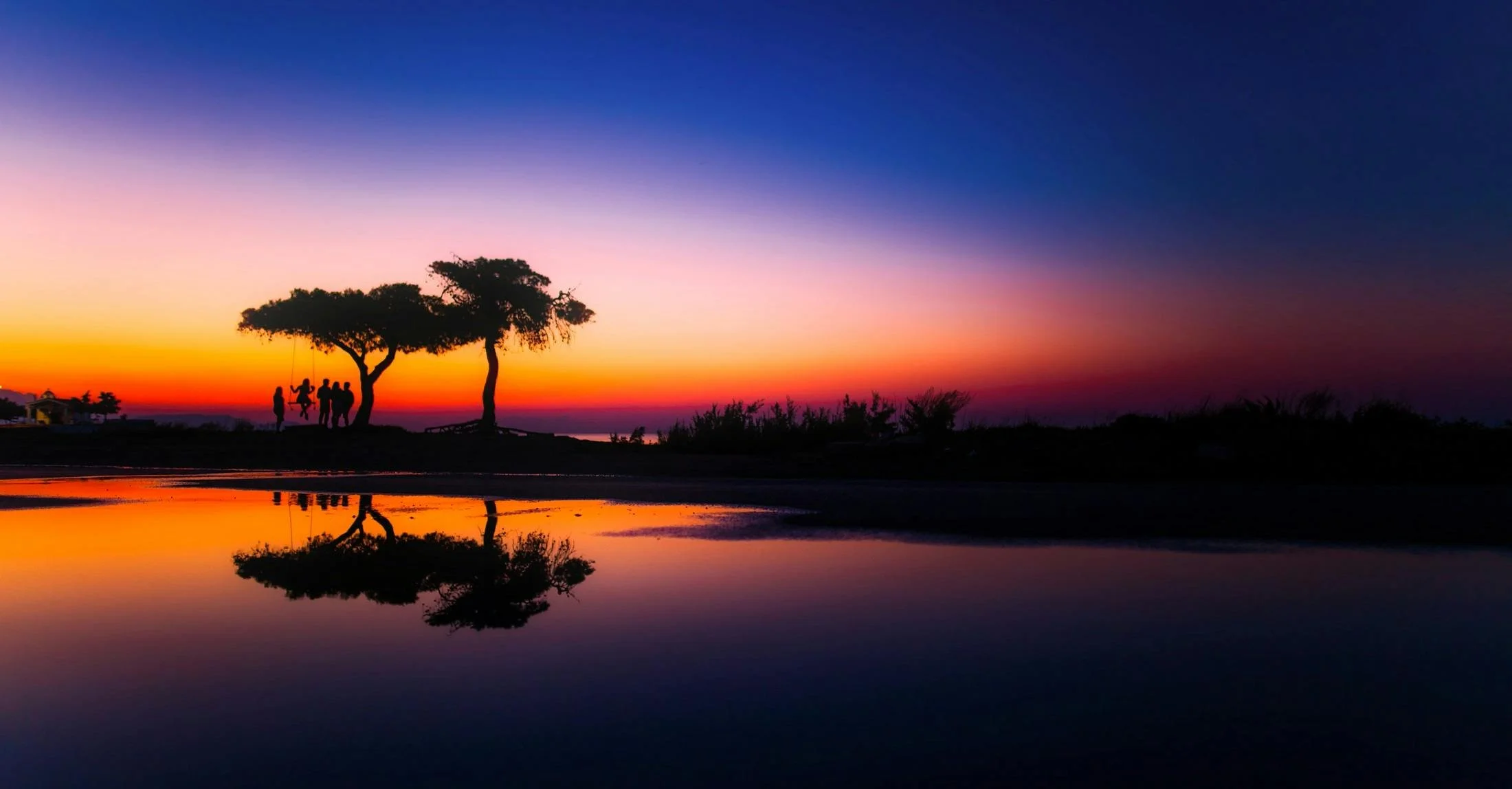 Silhouettes of a group of children playing on a swing and a tree with a colorful sunset sky reflected on water.