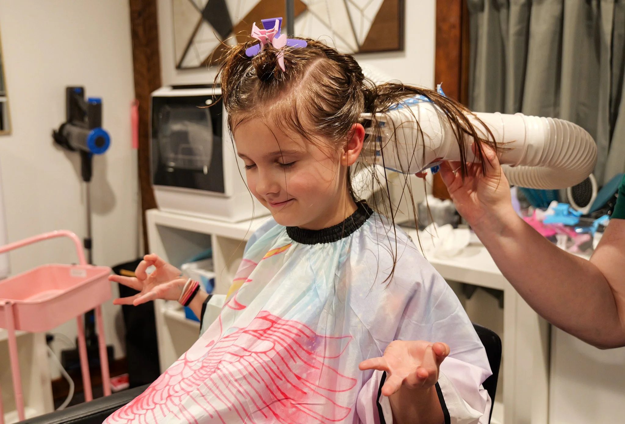 A girl sitting in a salon chair getting her hair dried by a hair dryer held by a stylist. The girl has wet hair and is wearing a colorful cape.