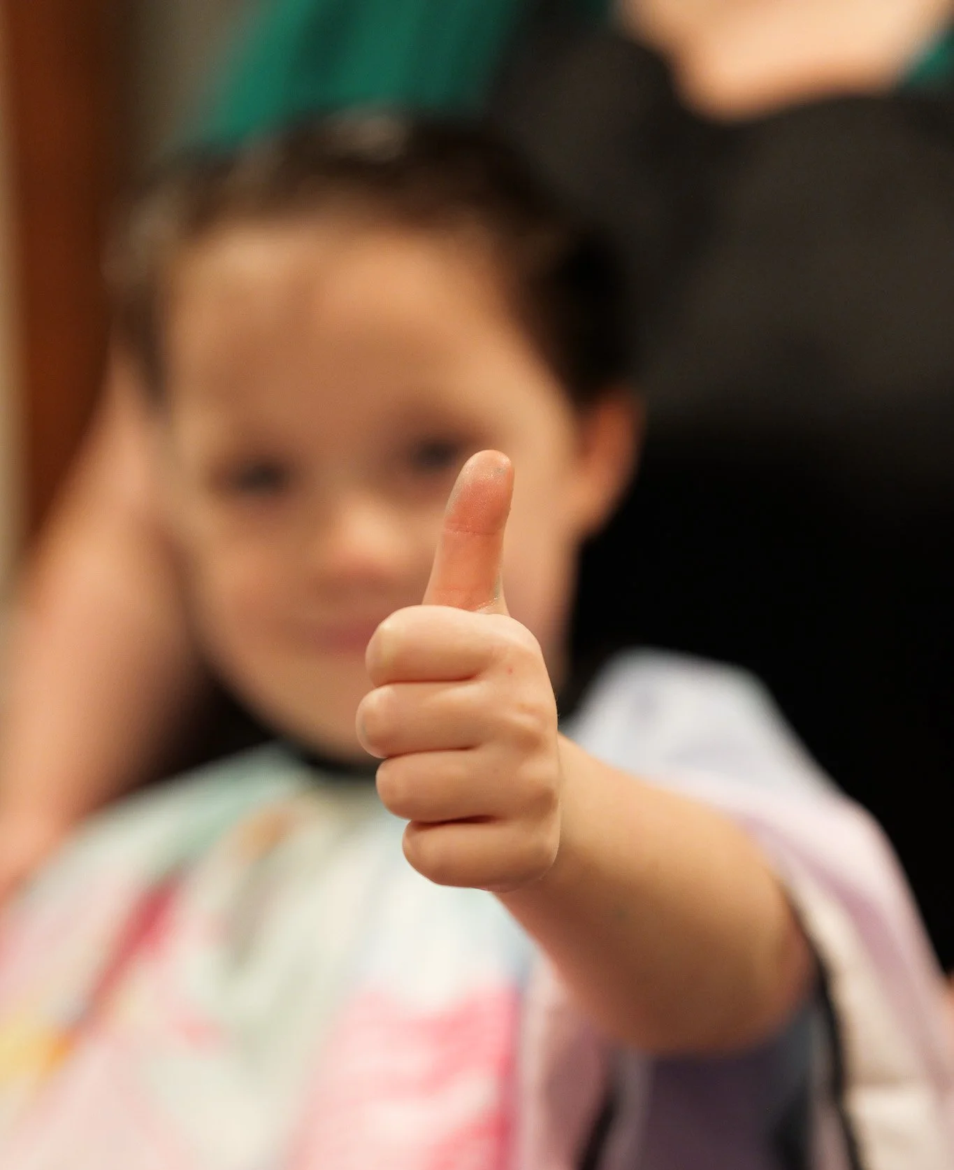 A young child with a blurred face giving a thumbs-up gesture, wearing a colorful shirt and a green cap.