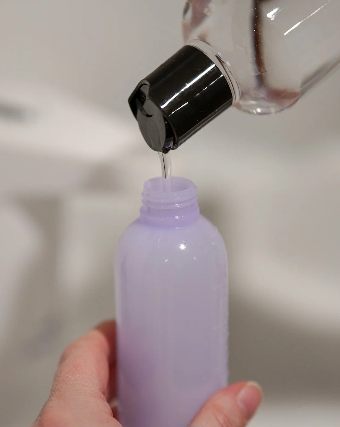 A person pouring a clear liquid from a bottle into a purple plastic bottle.