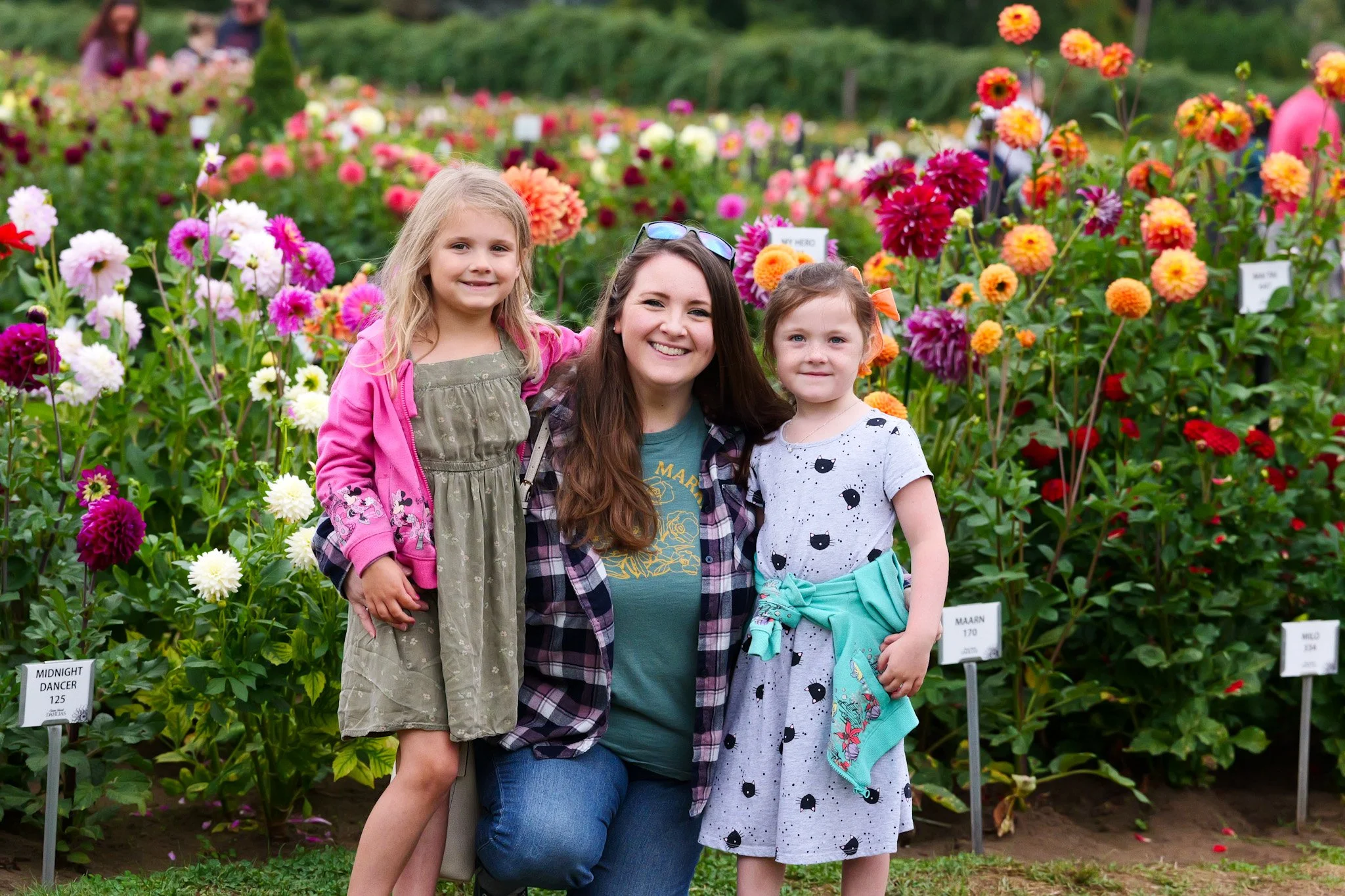 A woman with long brown hair and two young girls smiling in a colorful flower garden.