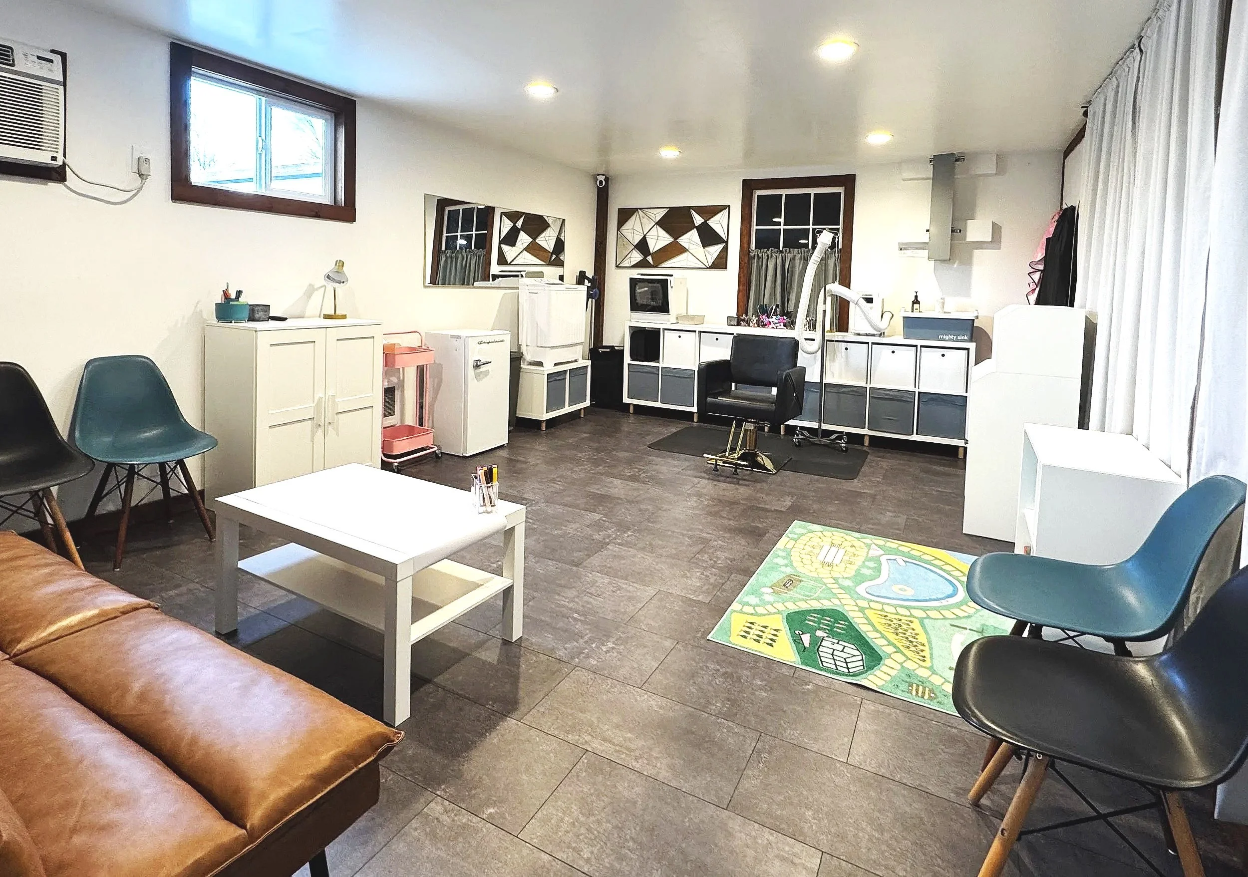 A tidy room with a brown leather sofa, a white coffee table, and multiple chairs including blue and black ones. There is a small colorful rug on the floor. The back of the room has white cabinets, a computer monitor, and a chair in front of a work area. Windows with white curtains and a small window air conditioning unit are on the left. Overhead lighting illuminates the room.